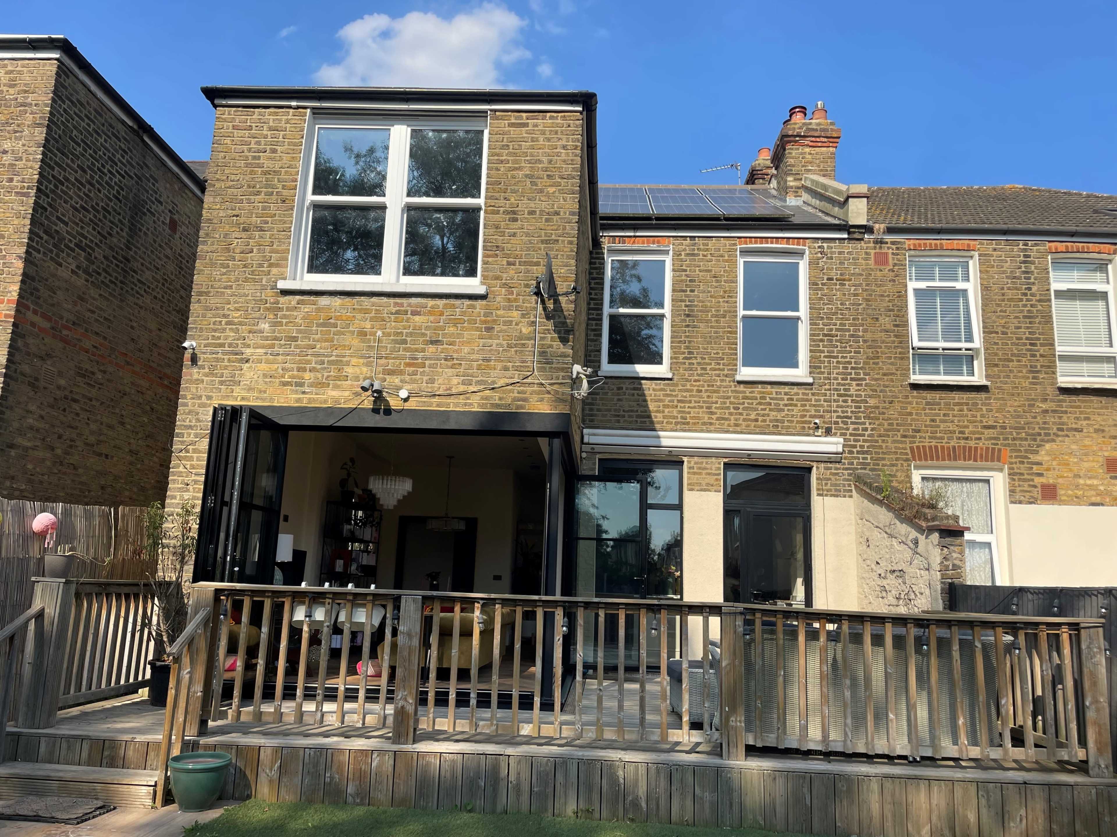 A two-story brick house with large windows, a wooden deck in the foreground, and solar panels on the roof, all set against a clear blue sky.