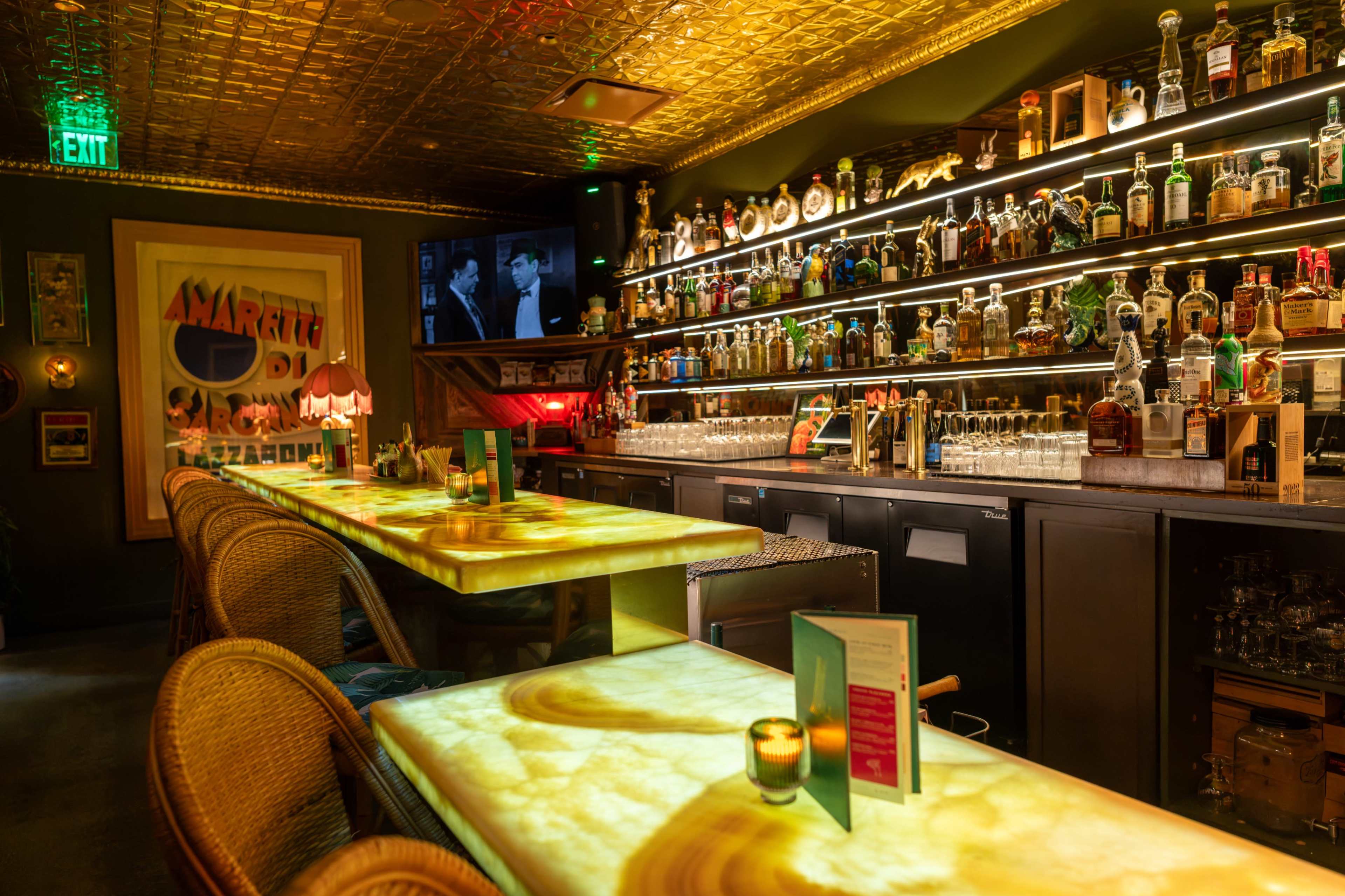 The image shows a stylish bar with illuminated, onyx-like tabletops, a wide selection of bottles on shelves behind the counter, and a vintage television display.