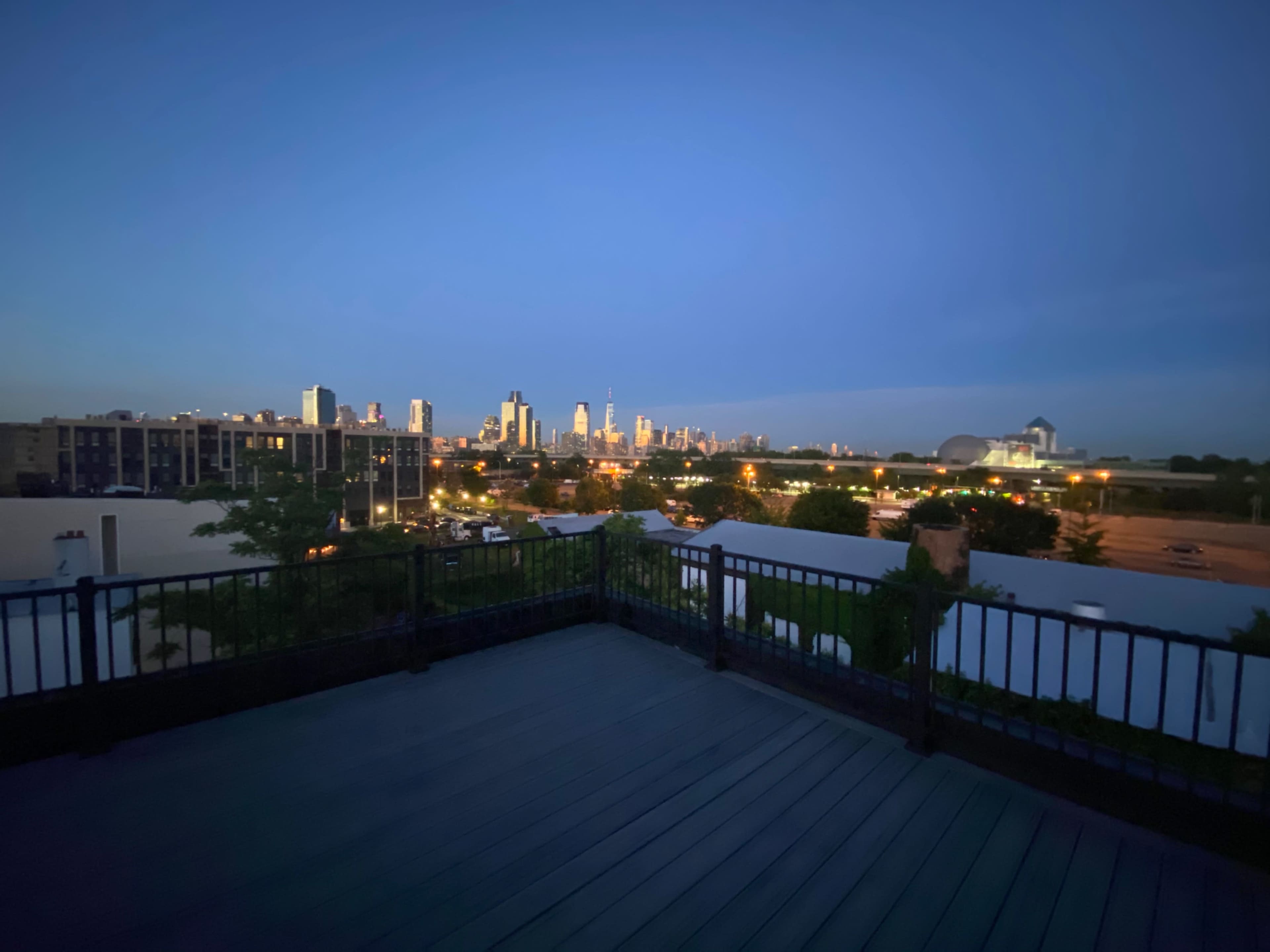 A city skyline is visible at dusk, with buildings illuminated against the darkening sky and a rooftop balcony in the foreground.
