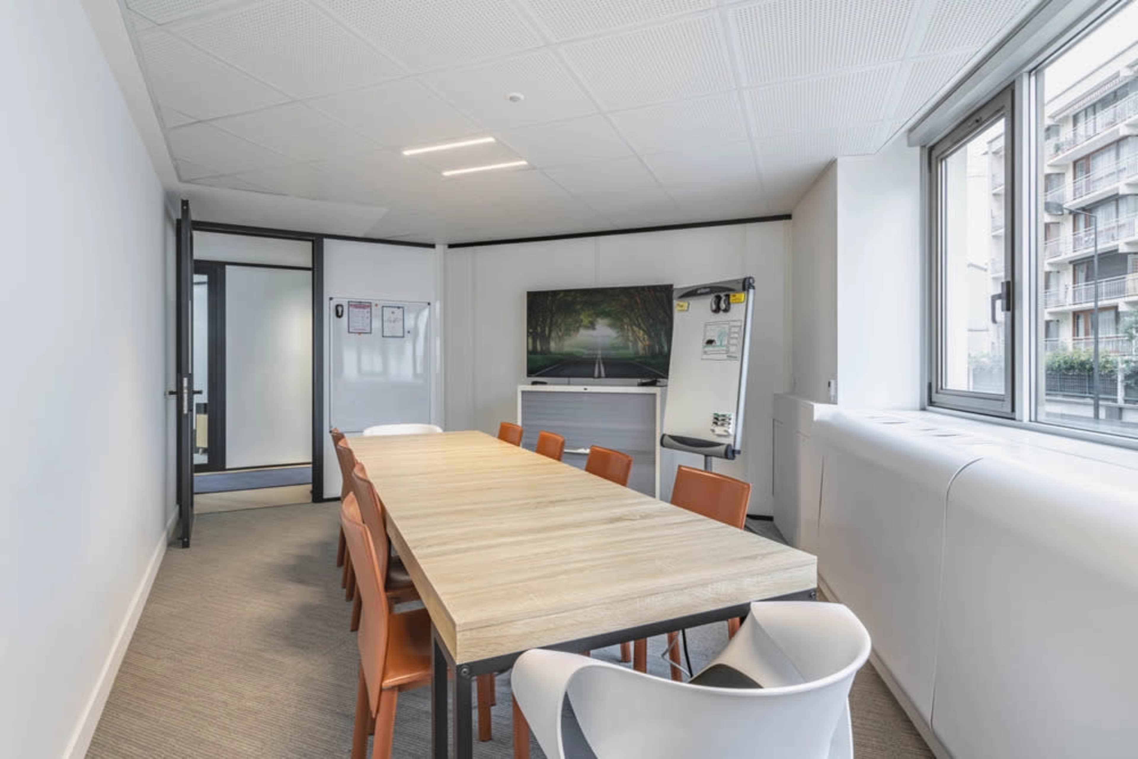 The image shows a modern meeting room featuring a long wooden table surrounded by orange and white chairs, with a large window allowing natural light and a picture of a green pathway on the wall.