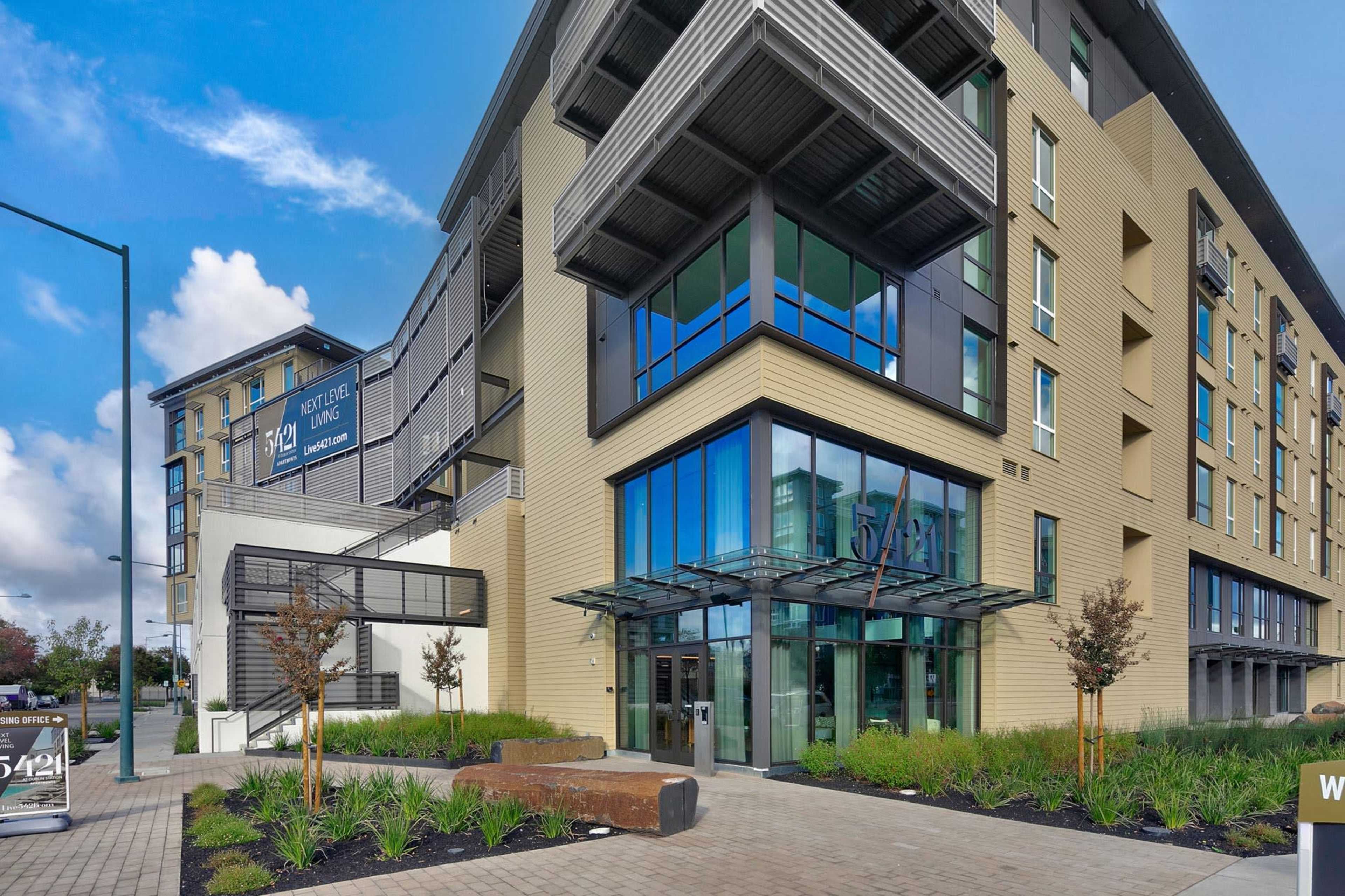 The image shows a modern multi-story building with large glass windows and a prominent entrance, surrounded by landscaping and a paved walkway.