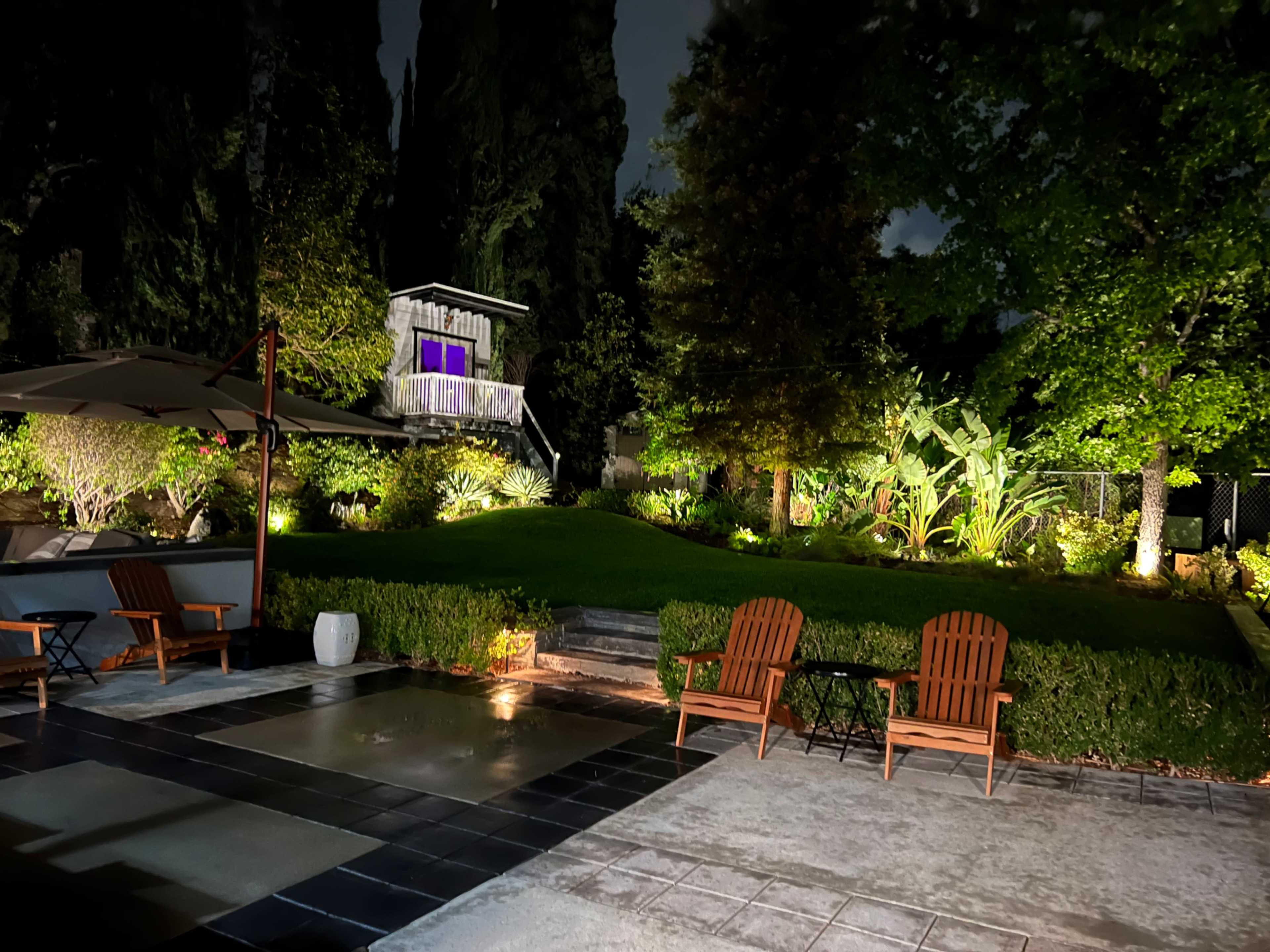 The image shows a well-lit backyard featuring two wooden chairs, a table, and a multi-level garden with various plants and trees under a night sky.