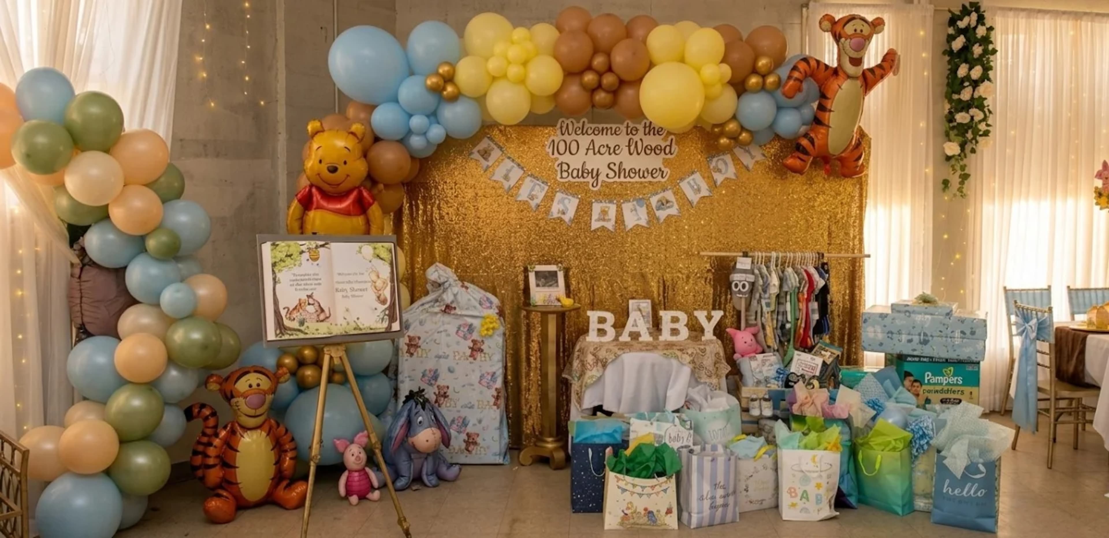 The image shows a decorated area for a baby shower, featuring a backdrop with a gold sequin curtain, balloon arrangements, and gifts on display.