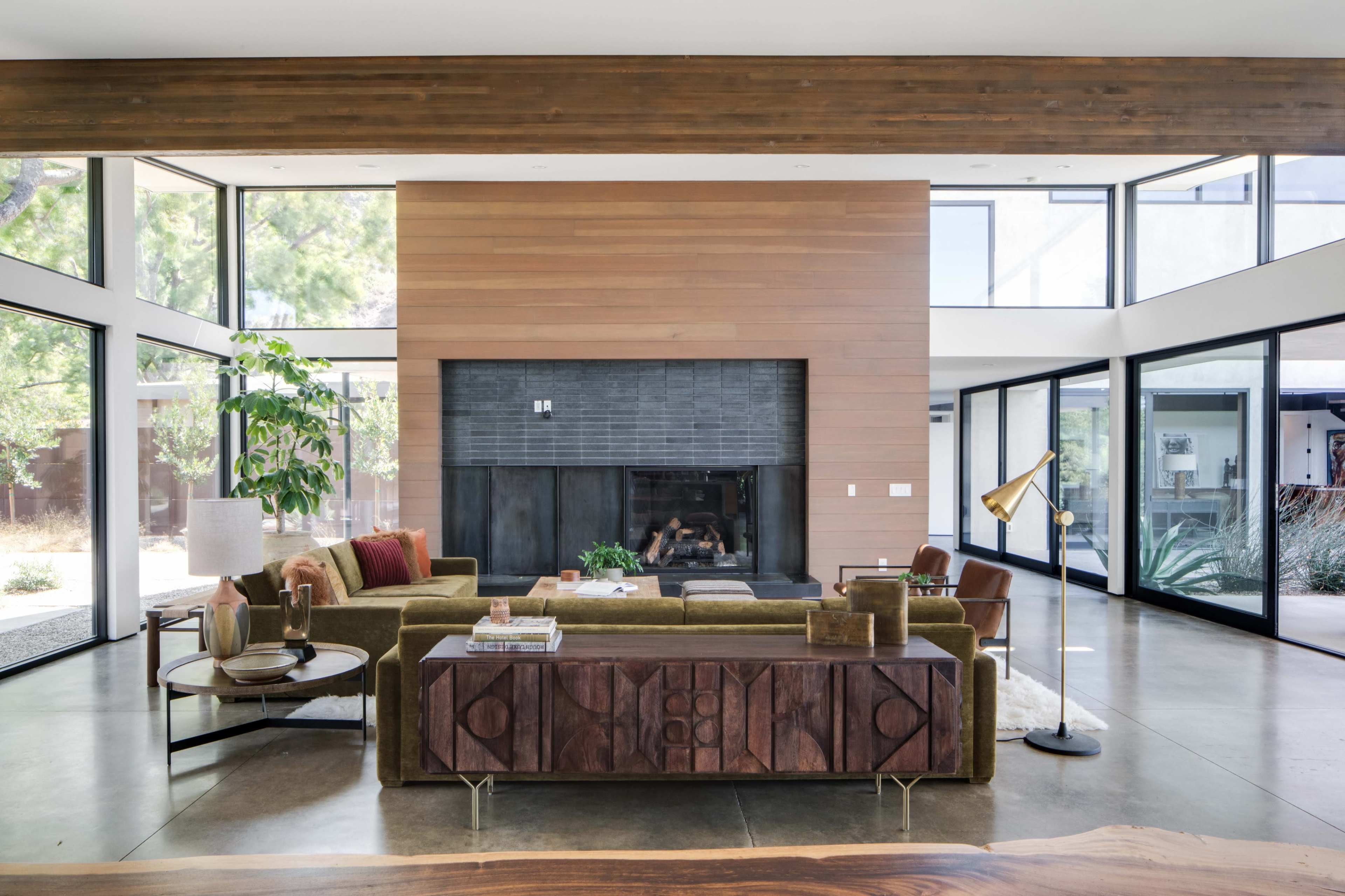The image shows a modern living room featuring a green sofa, wooden accent wall, large windows, and a fireplace.