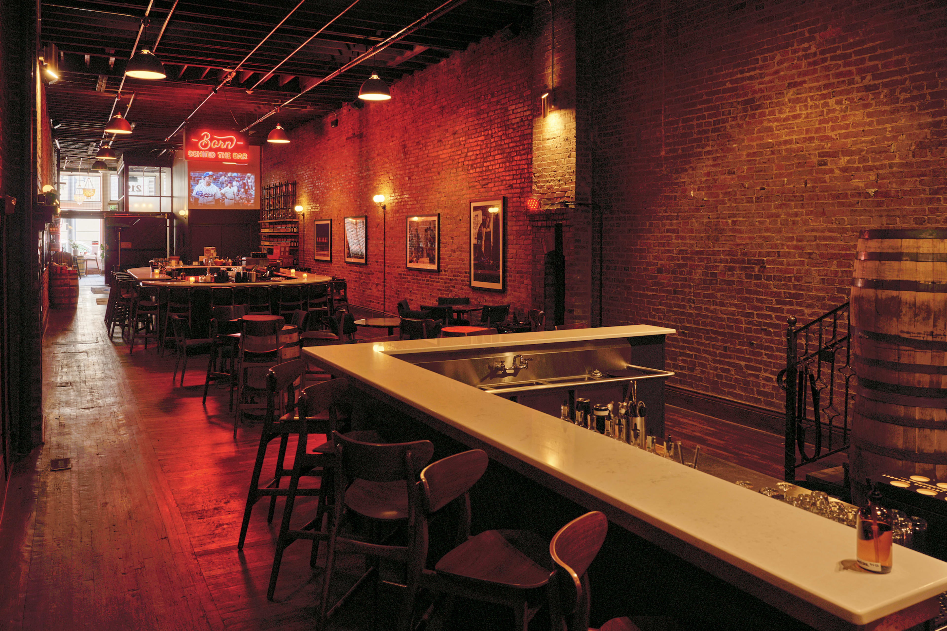 The interior of a rustic restaurant features exposed brick walls, wooden floors, and a long bar with seating, illuminated by a neon sign in the background.