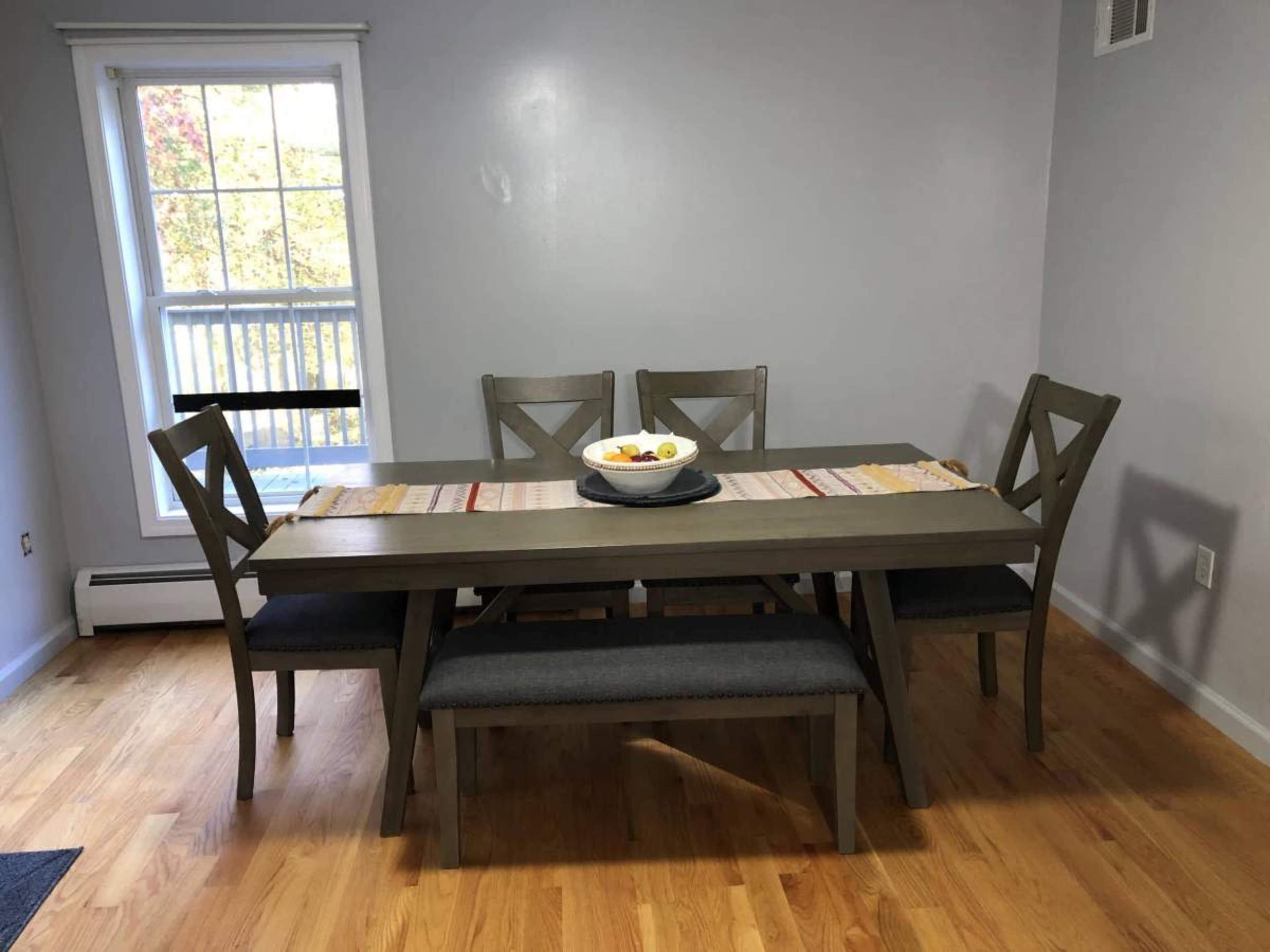 A wooden dining table with four chairs and a bench is set against a gray wall, with a bowl of fruit on the table and a window in the background.