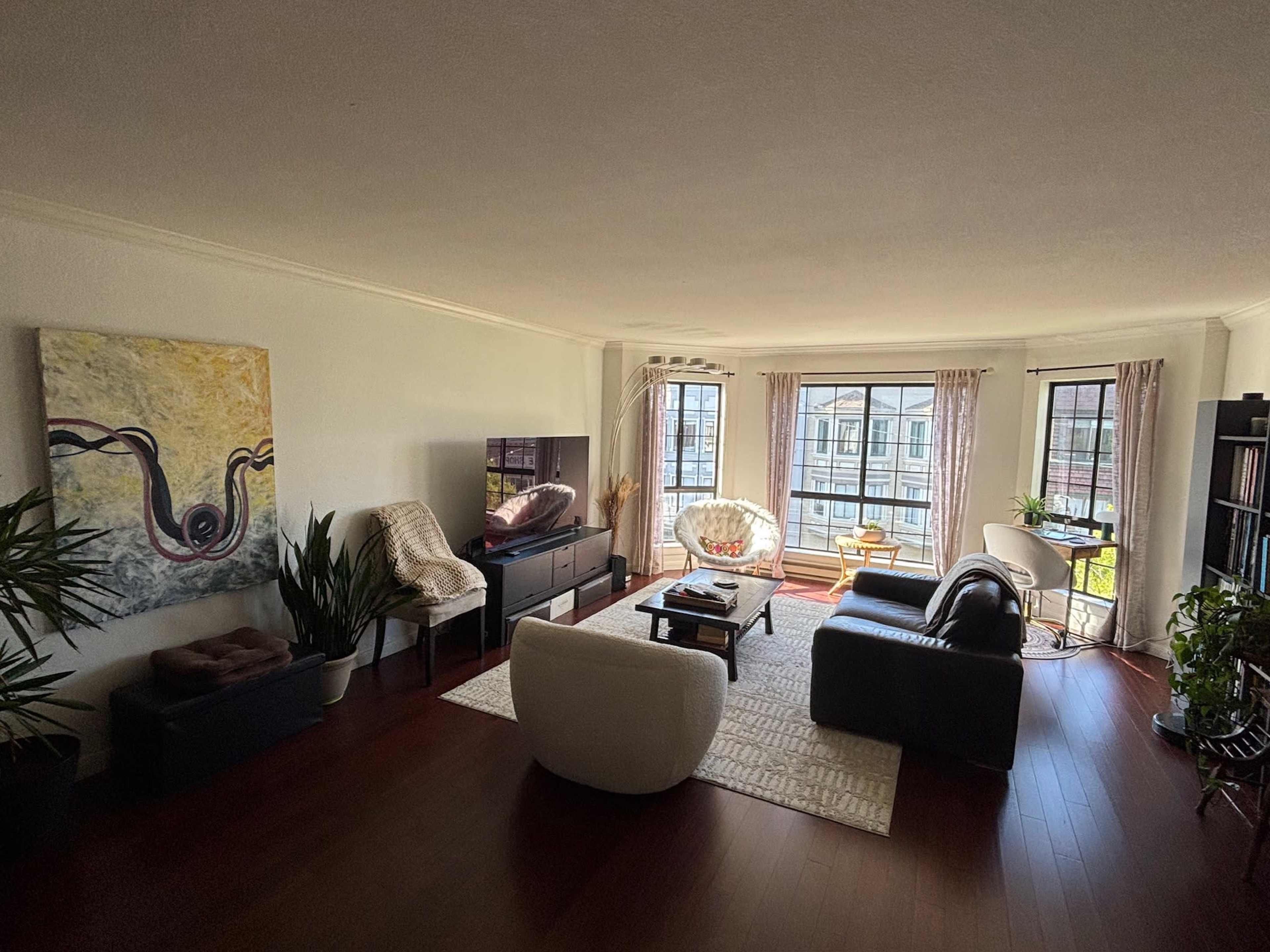 A modern living room features a mix of seating areas, a television, and large windows allowing natural light, with plants and artwork adding to the decor.