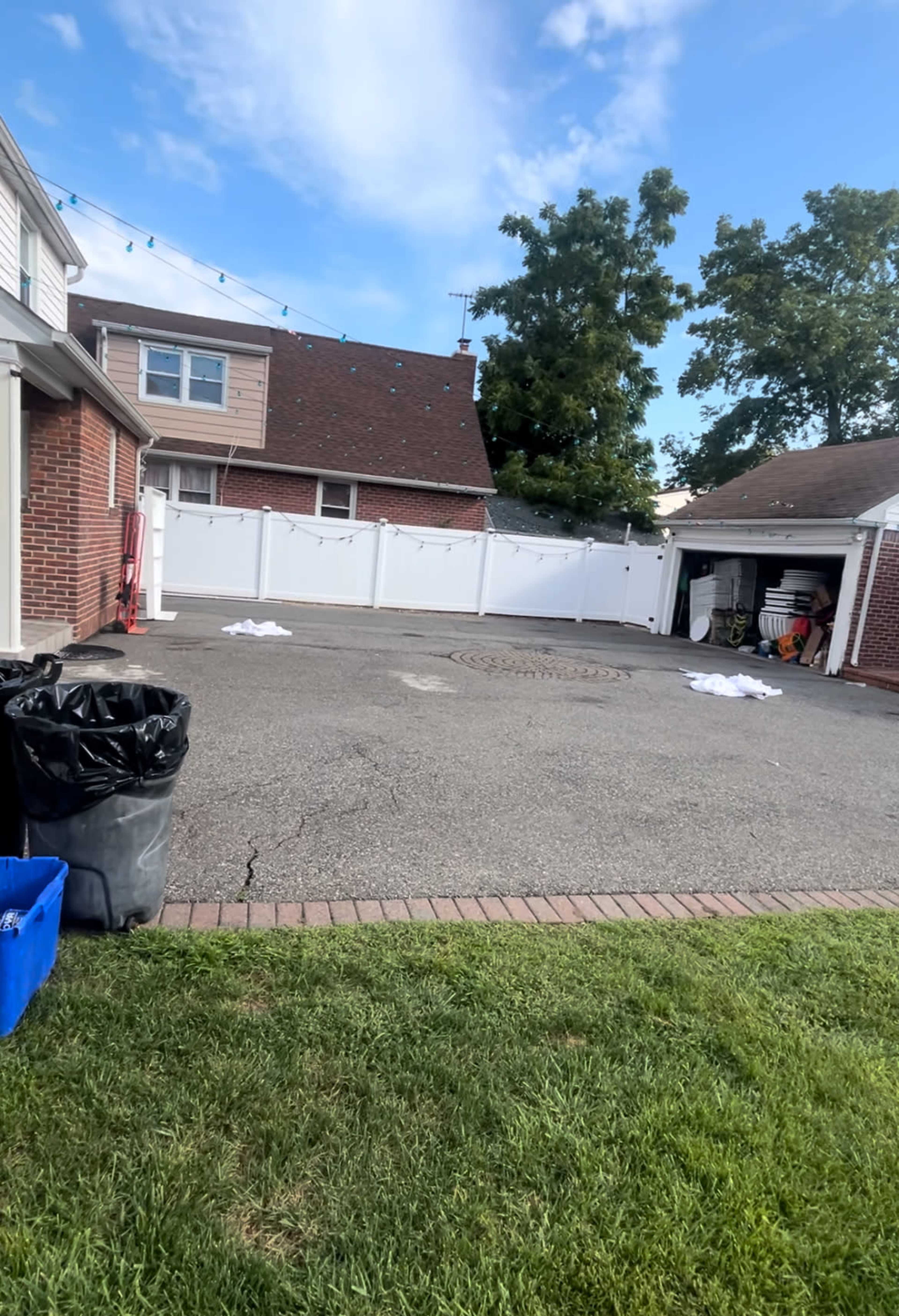 The image shows a paved backyard area with a garage to the right, a large garbage can in the foreground, and several white items laid out on the ground.