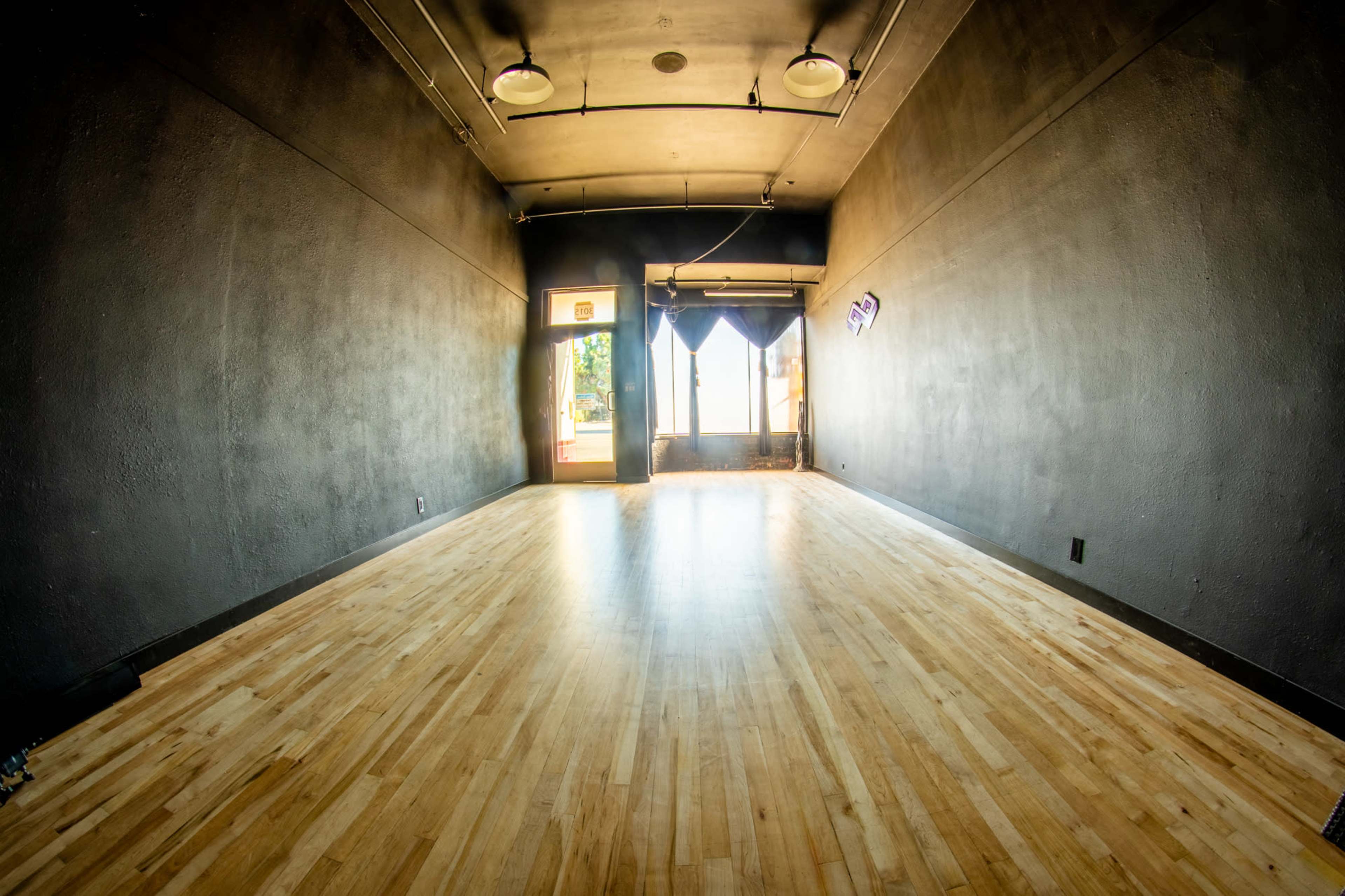 The image shows a vacant room with dark walls, a wooden floor, and a doorway at the far end that opens to natural light.
