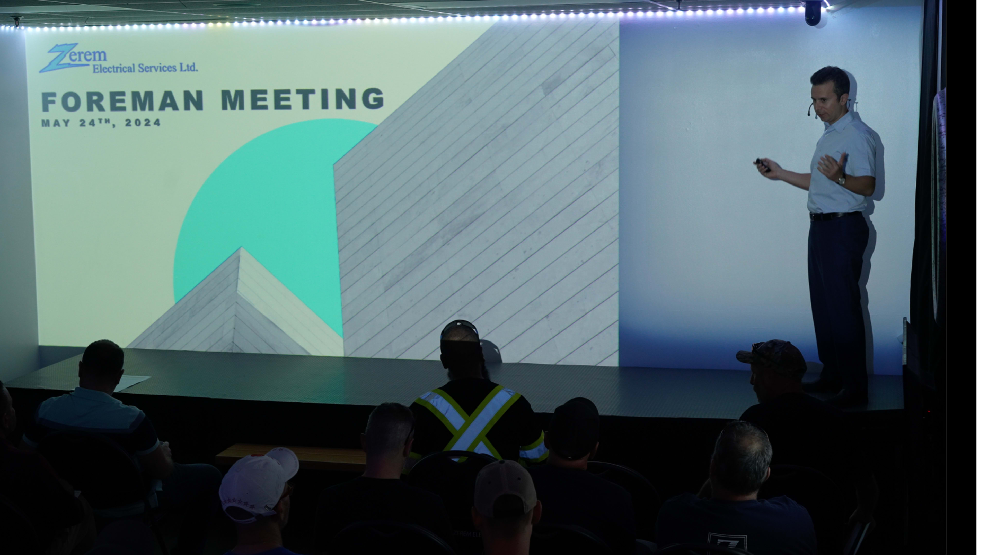 A presenter stands in front of a screen displaying "Foreman Meeting" along with the date May 24th, 2024, while an audience seated in semi-darkness listens attentively.