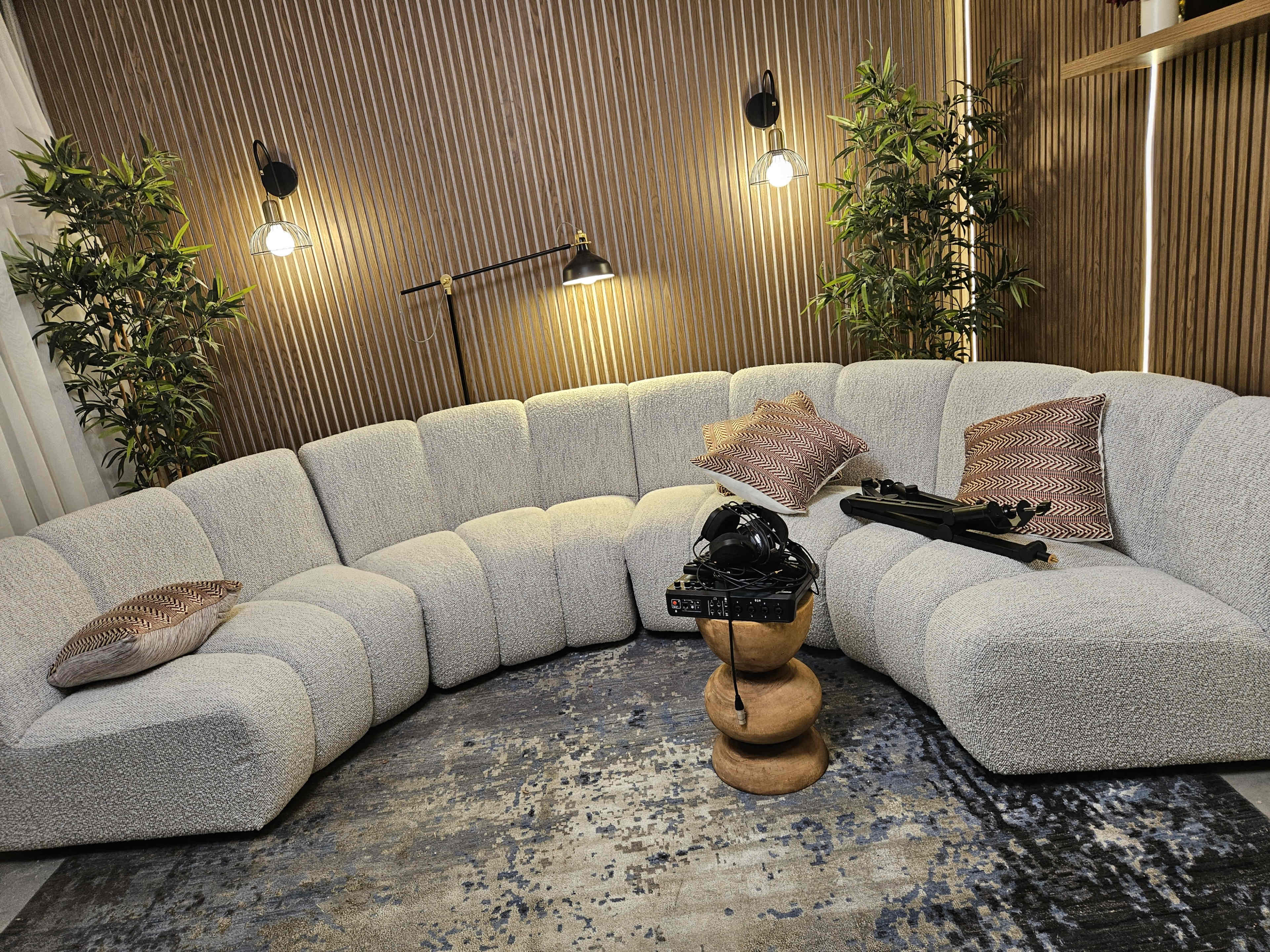 The image shows a curved, textured sofa positioned in a cozy room with warm lighting, surrounded by potted plants and a decorative rug.