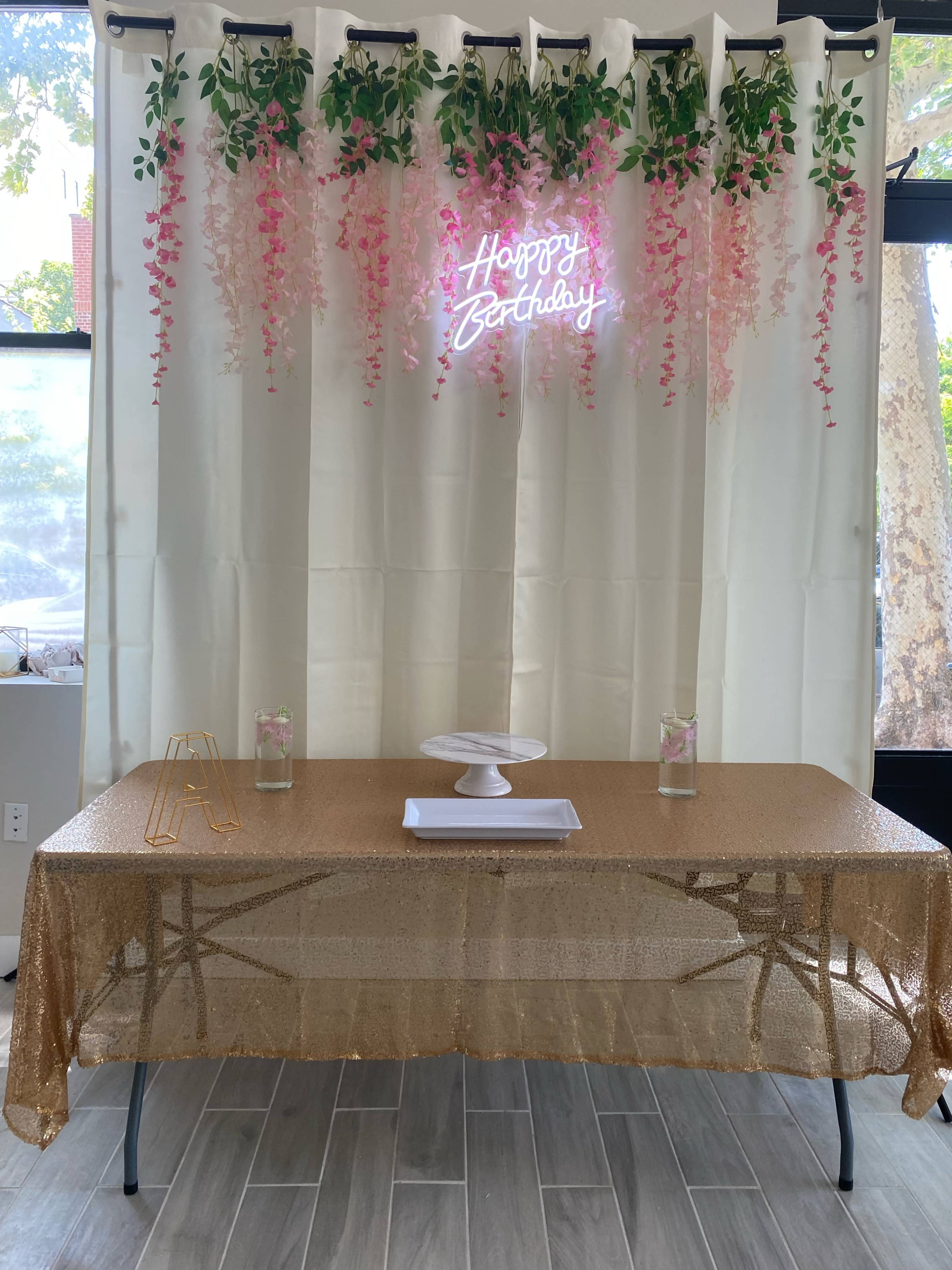 A table adorned with a gold sequin tablecloth is set in front of a white curtain decorated with hanging pink flowers and a neon sign that reads "Happy Birthday."