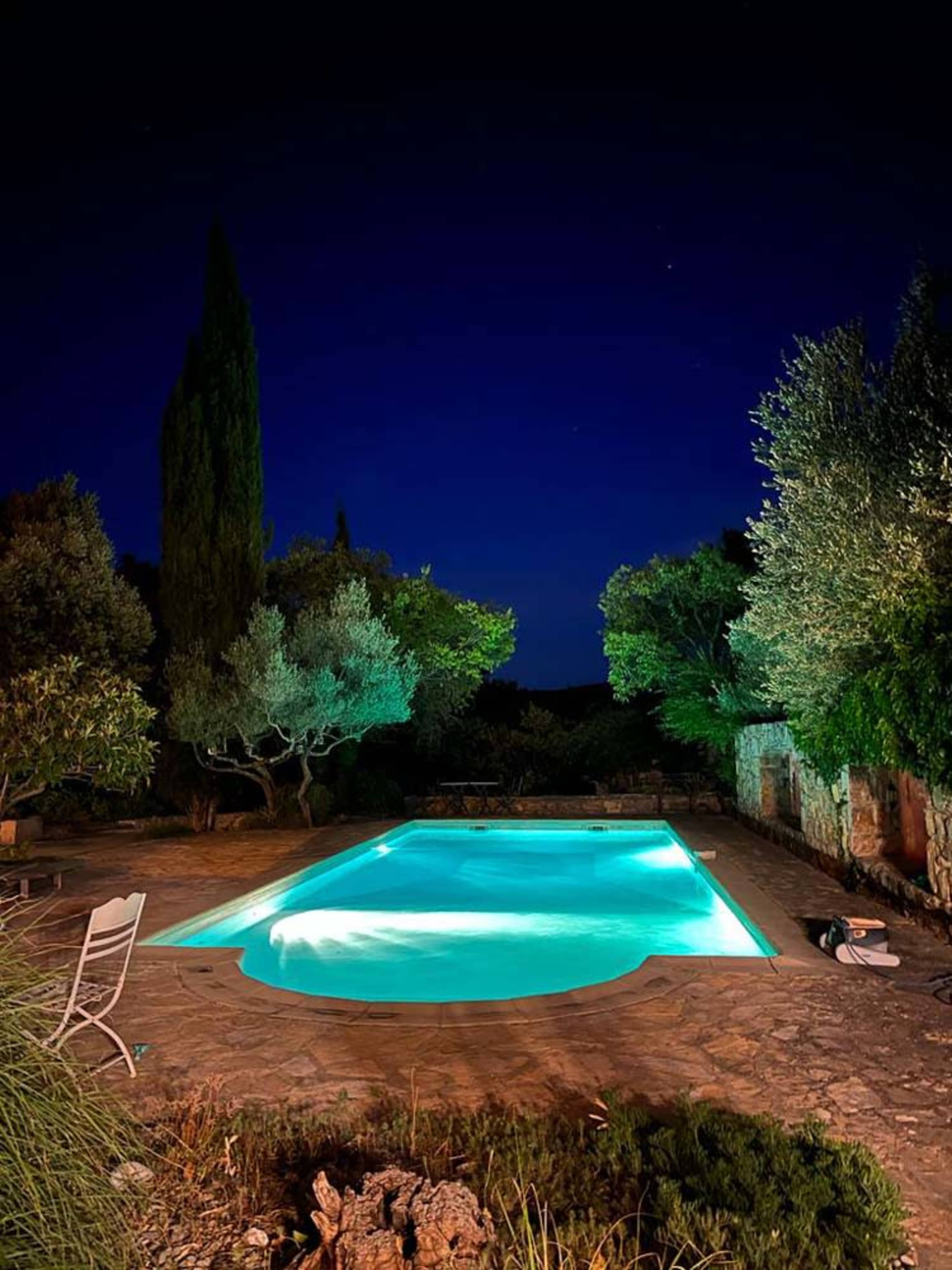 A brightly lit swimming pool is surrounded by trees and stone walls under a night sky.