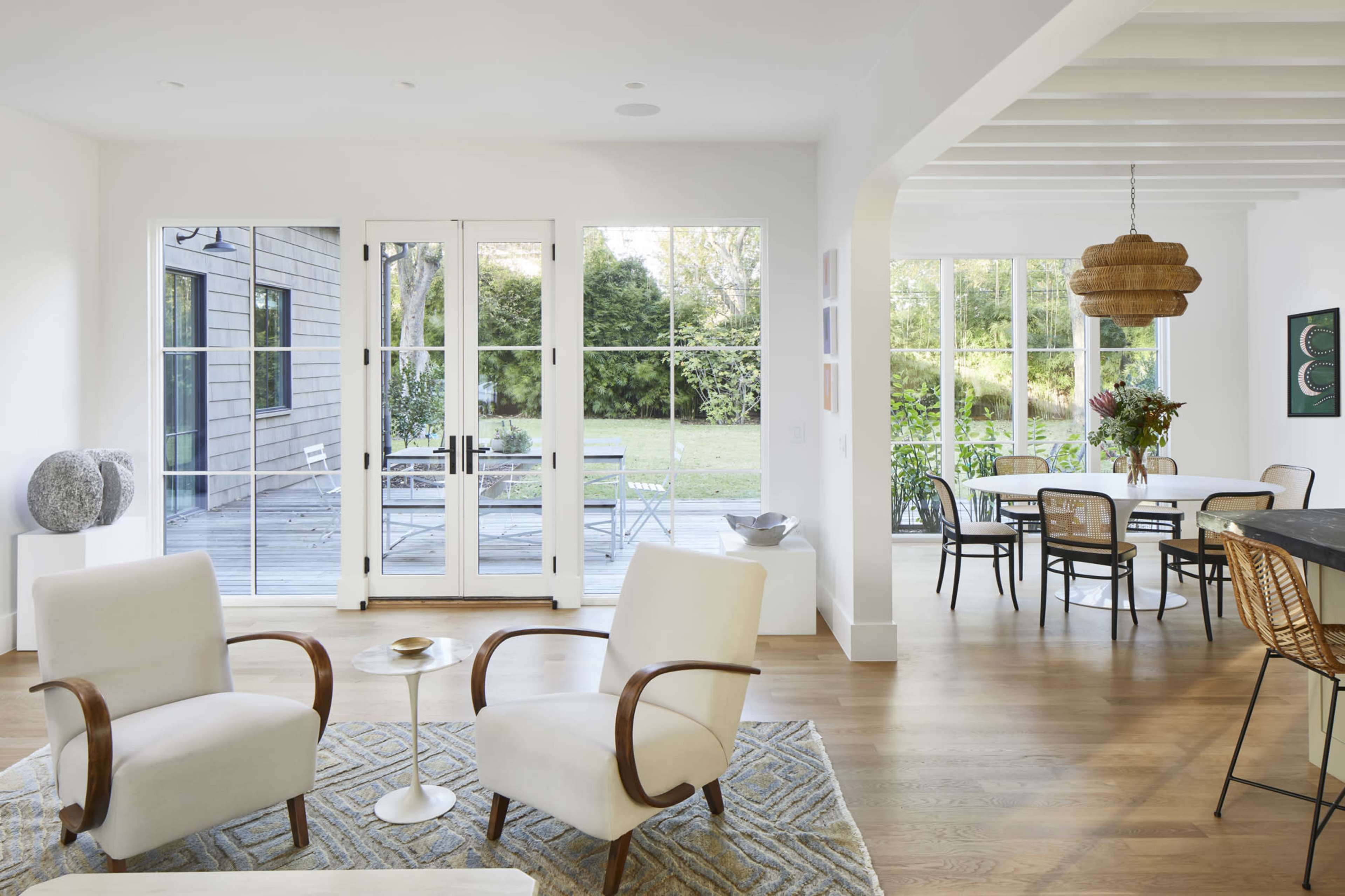 A bright, modern living space with two armchairs facing a small table, large glass doors leading to an outdoor area, and a dining table in the background.