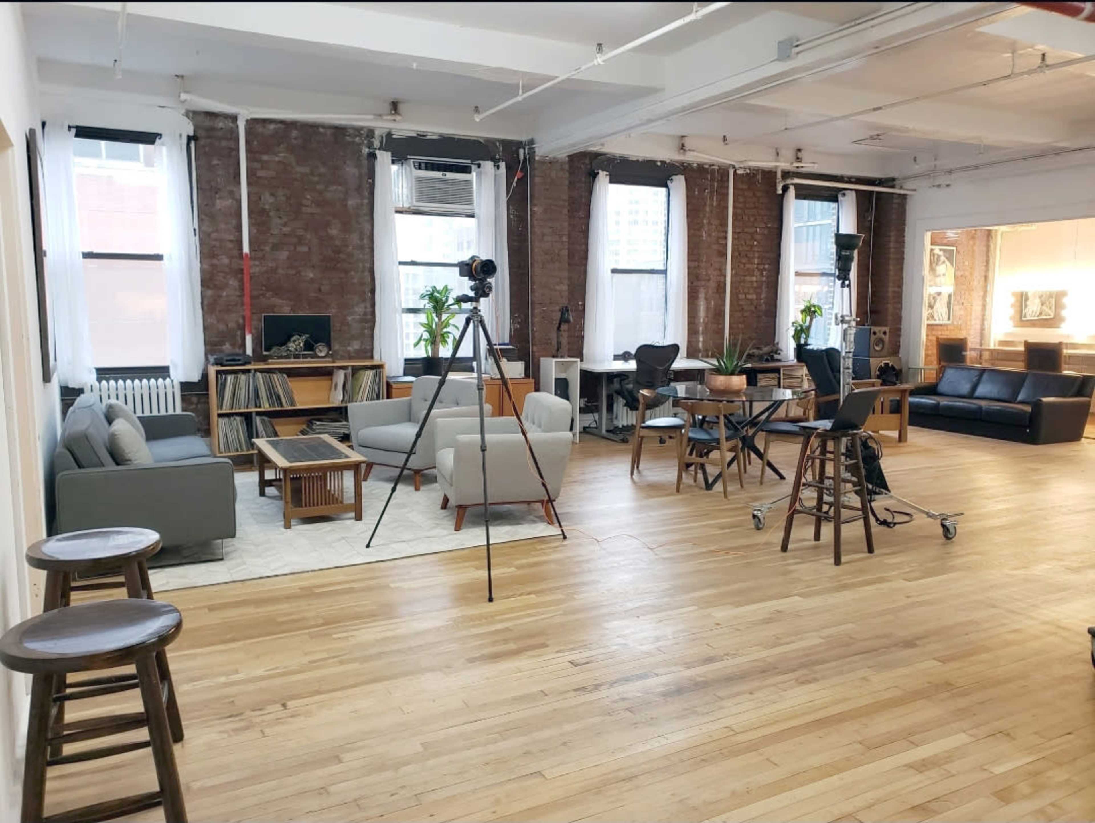 The image shows a spacious, well-lit room with hardwood floors, featuring various seating areas, a record player, and equipment set up for a photo shoot.