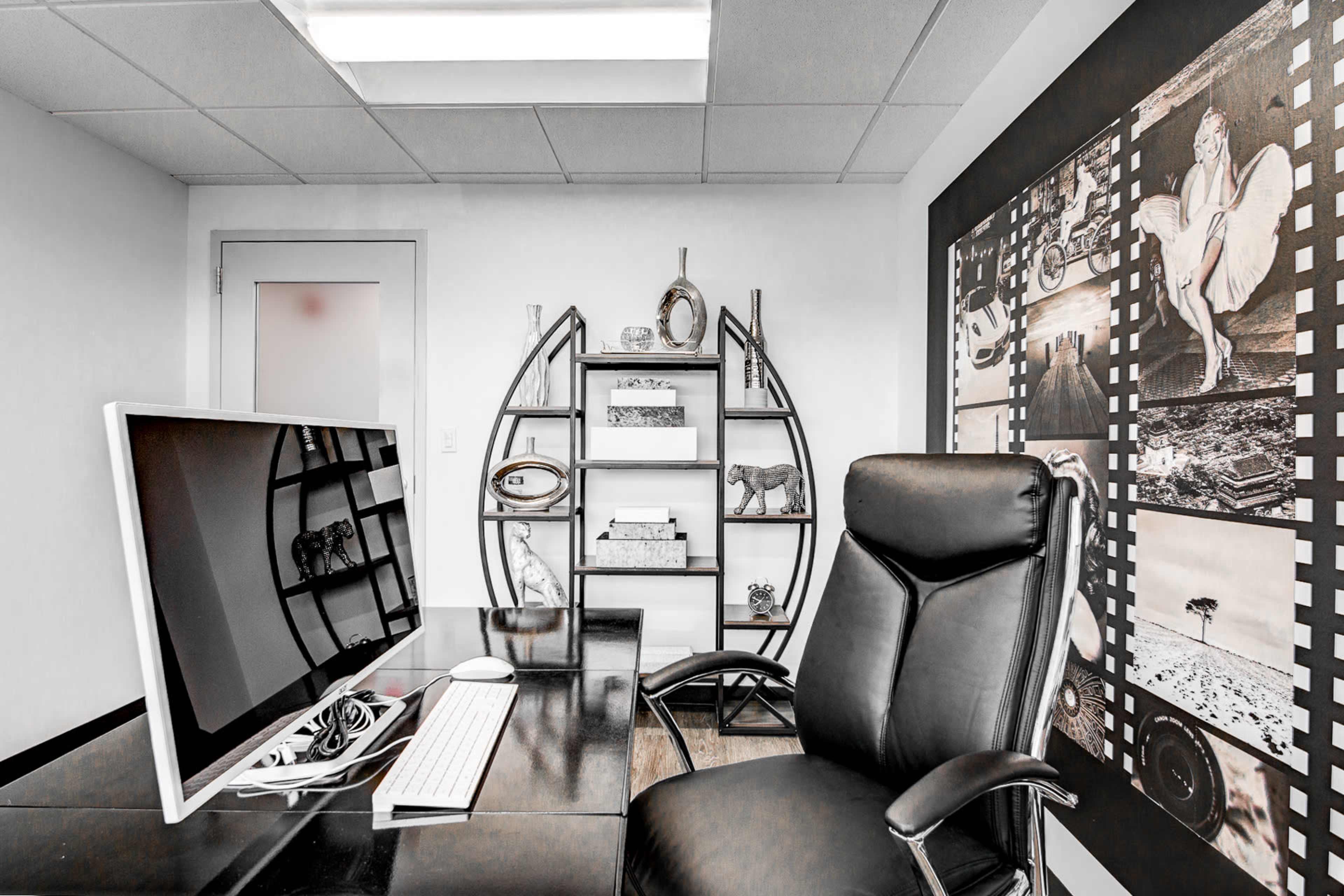A modern office space featuring a black desk with a computer, a black ergonomic chair, and a wall decorated with framed photos and a circular shelf.