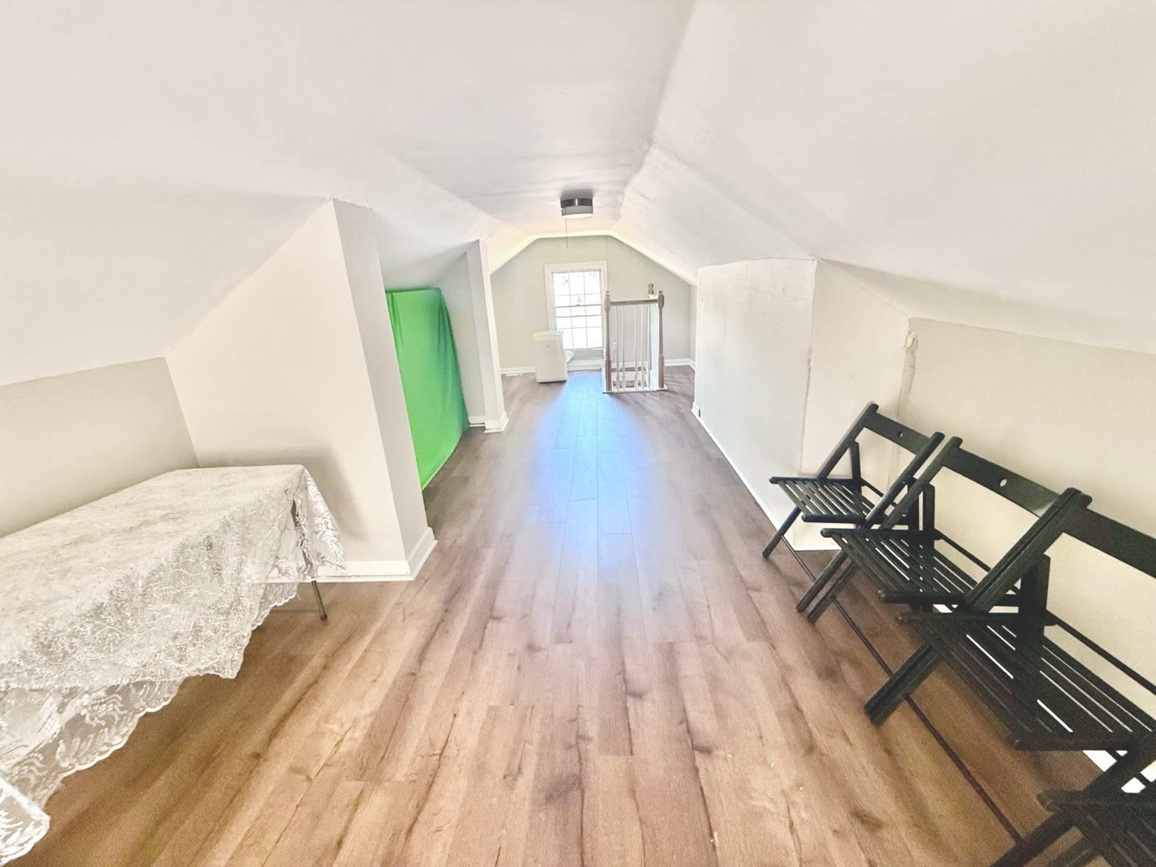 The image shows a narrow attic space with light wood flooring, a table covered with a lace cloth on one side, and several black folding chairs arranged along the wall.