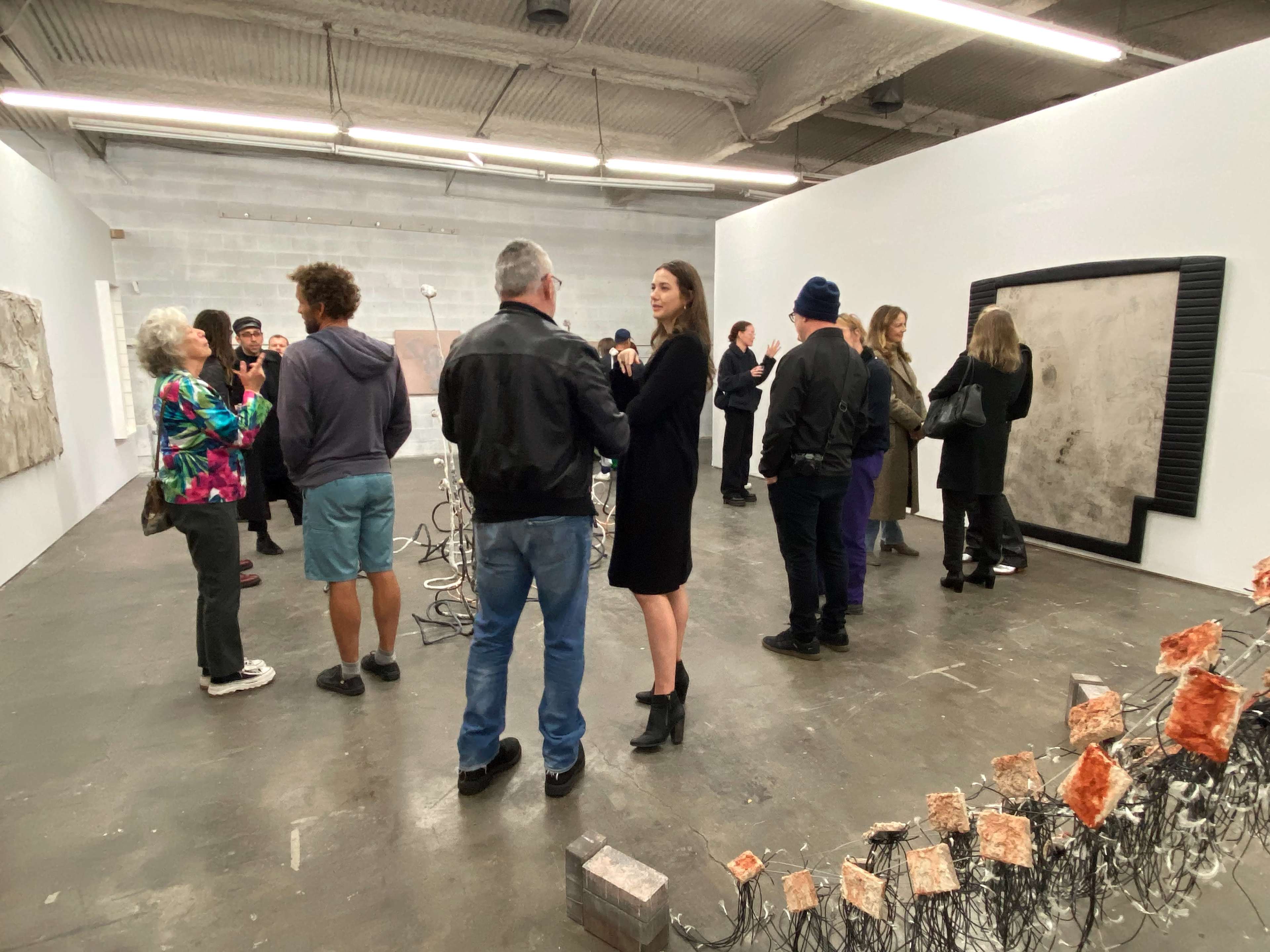A group of people discusses artworks in a gallery featuring abstract pieces and a sculptural installation.