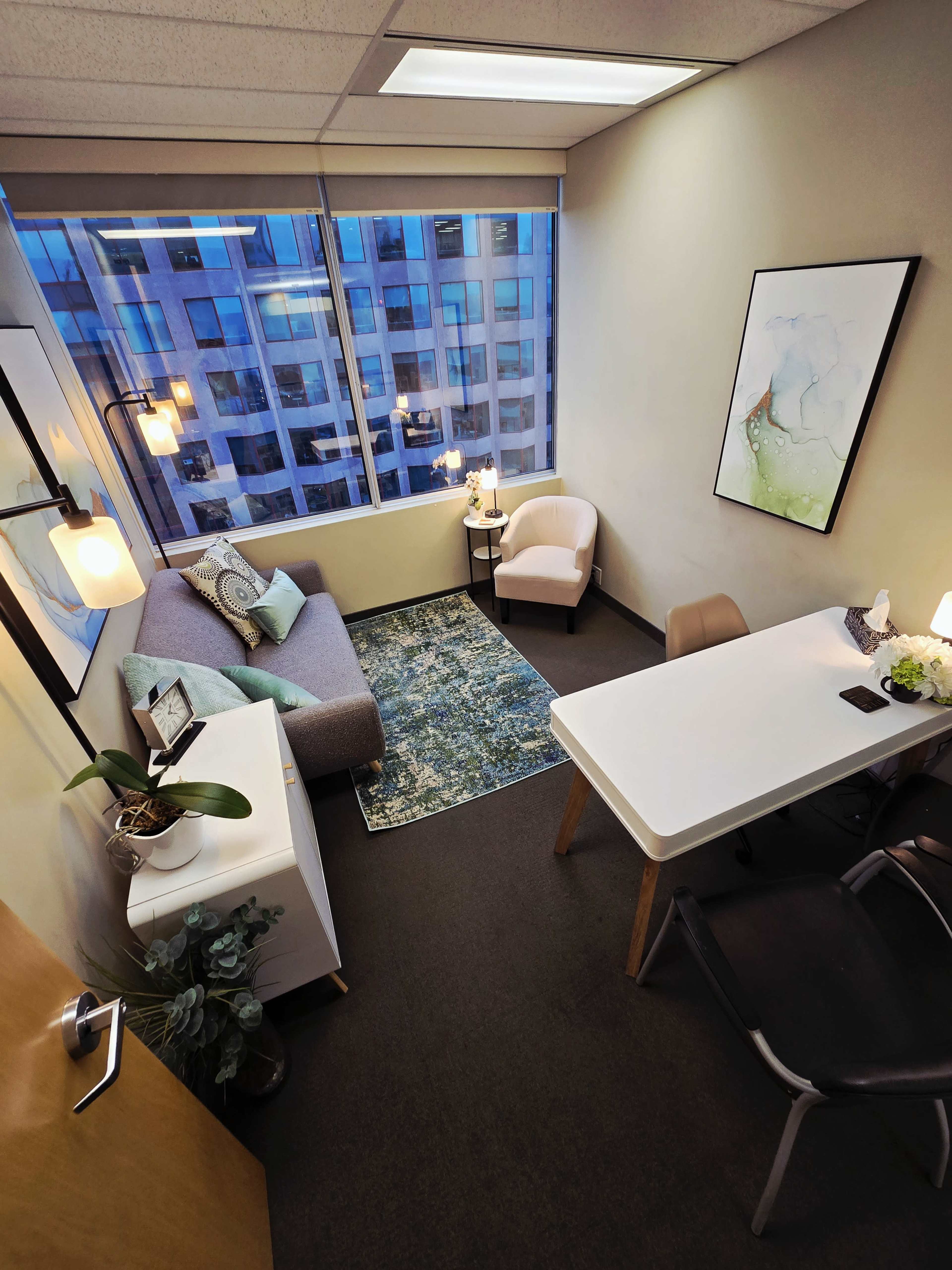 A small office features a gray sofa, a white desk, and a large window showcasing a cityscape, with decorative lighting and a blue area rug.