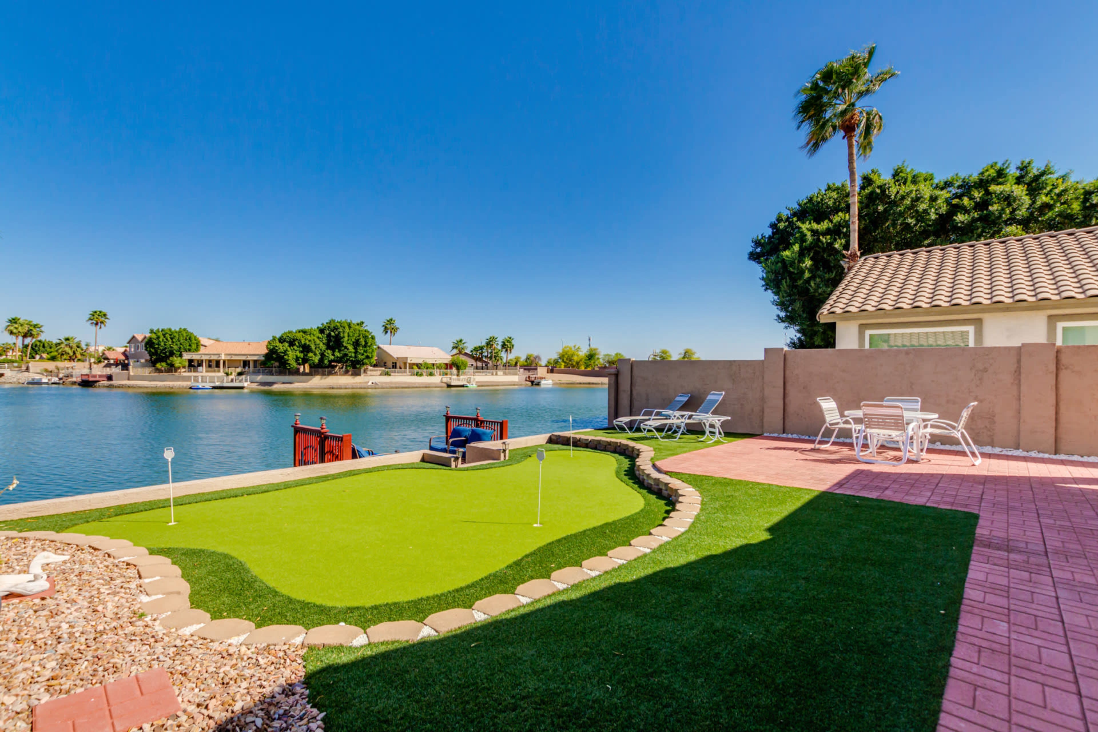A backyard features a putting green, patio furniture, and a view of the water and houses across the canal.