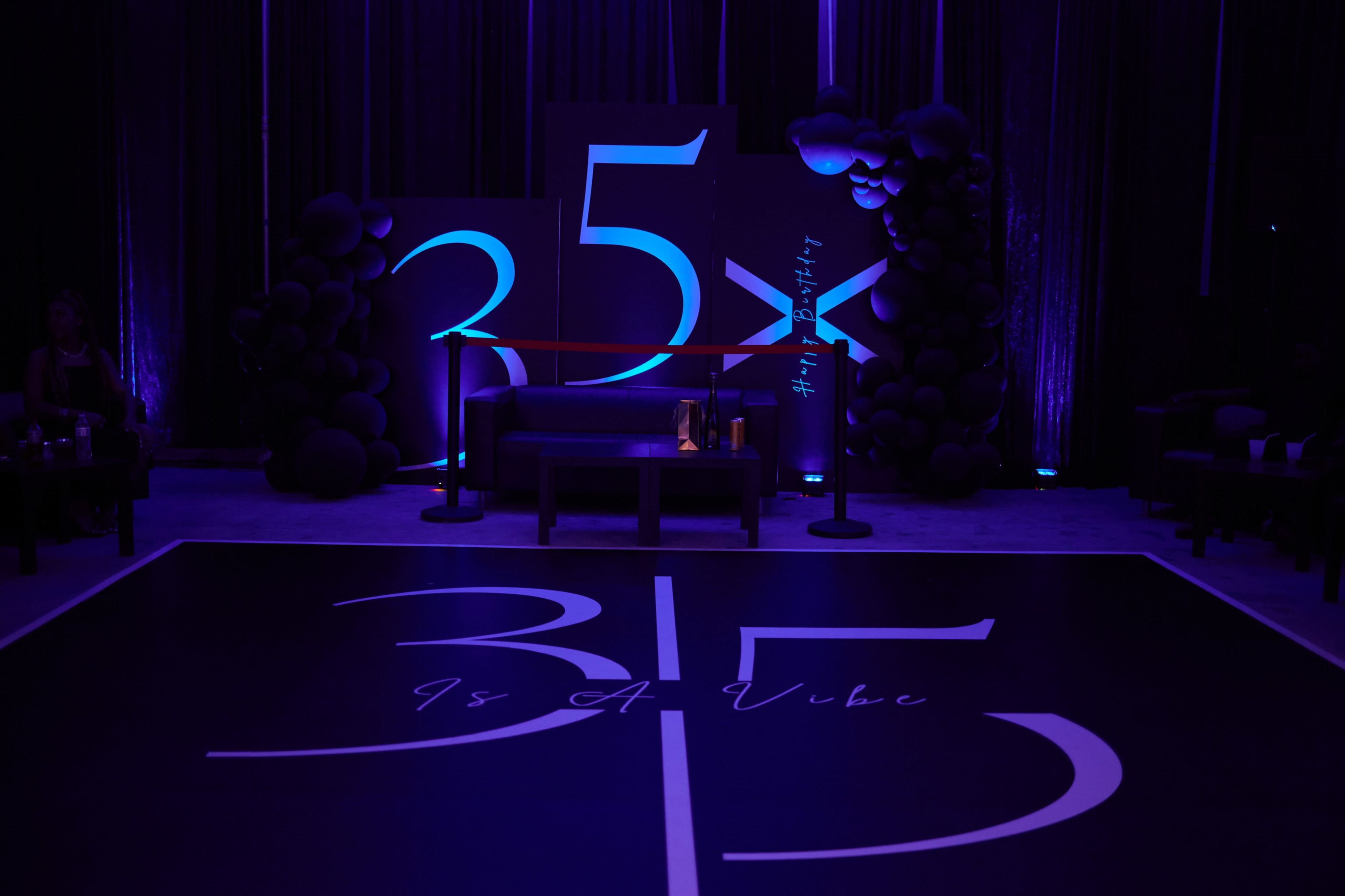 The image shows a stylish event space with a black and purple theme, featuring the number "35" prominently displayed on the floor and walls, along with decorative elements like balloons and a seating area.