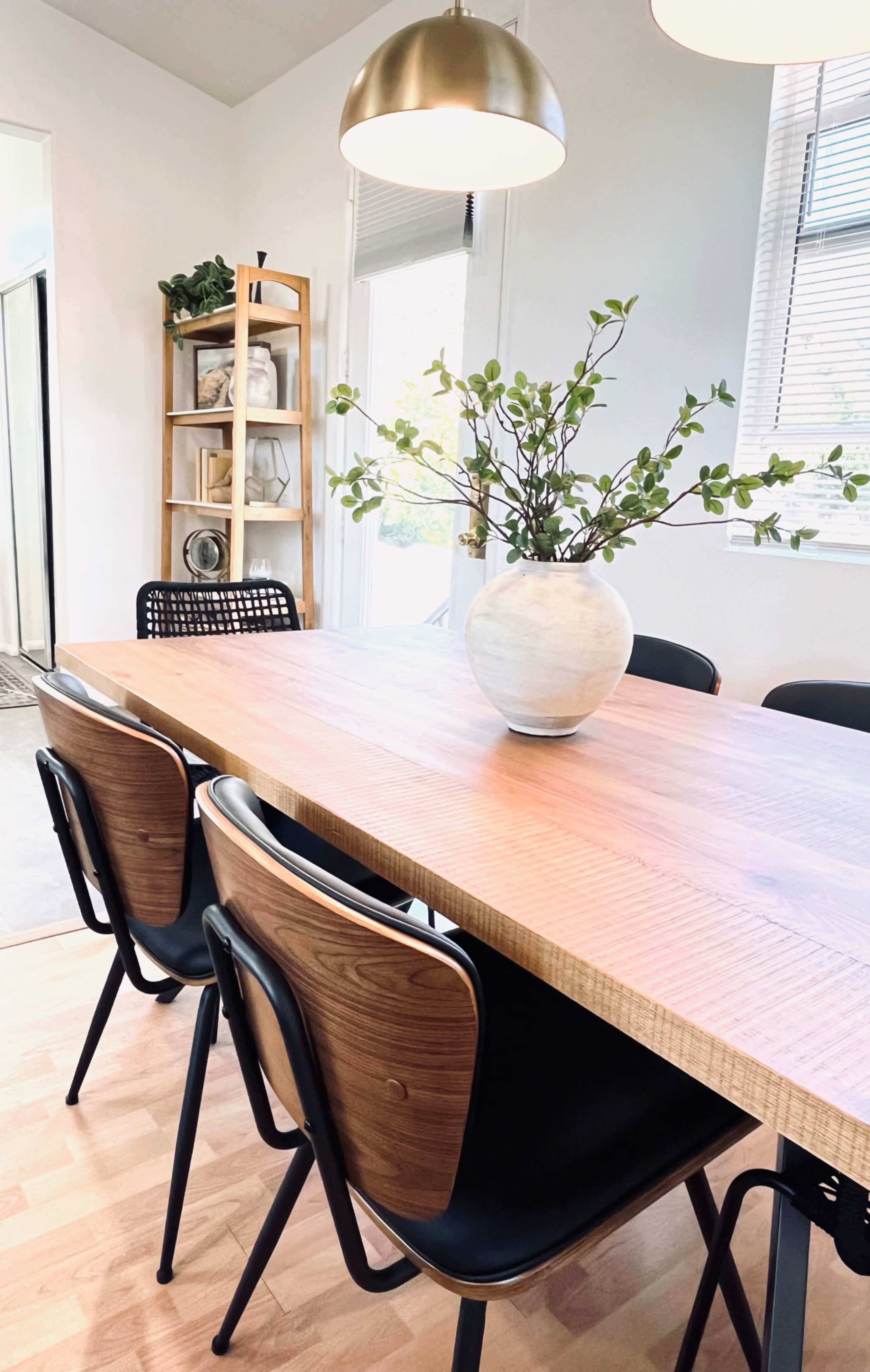 A wooden dining table is set with a vase of greenery, surrounded by black chairs in a well-lit room.