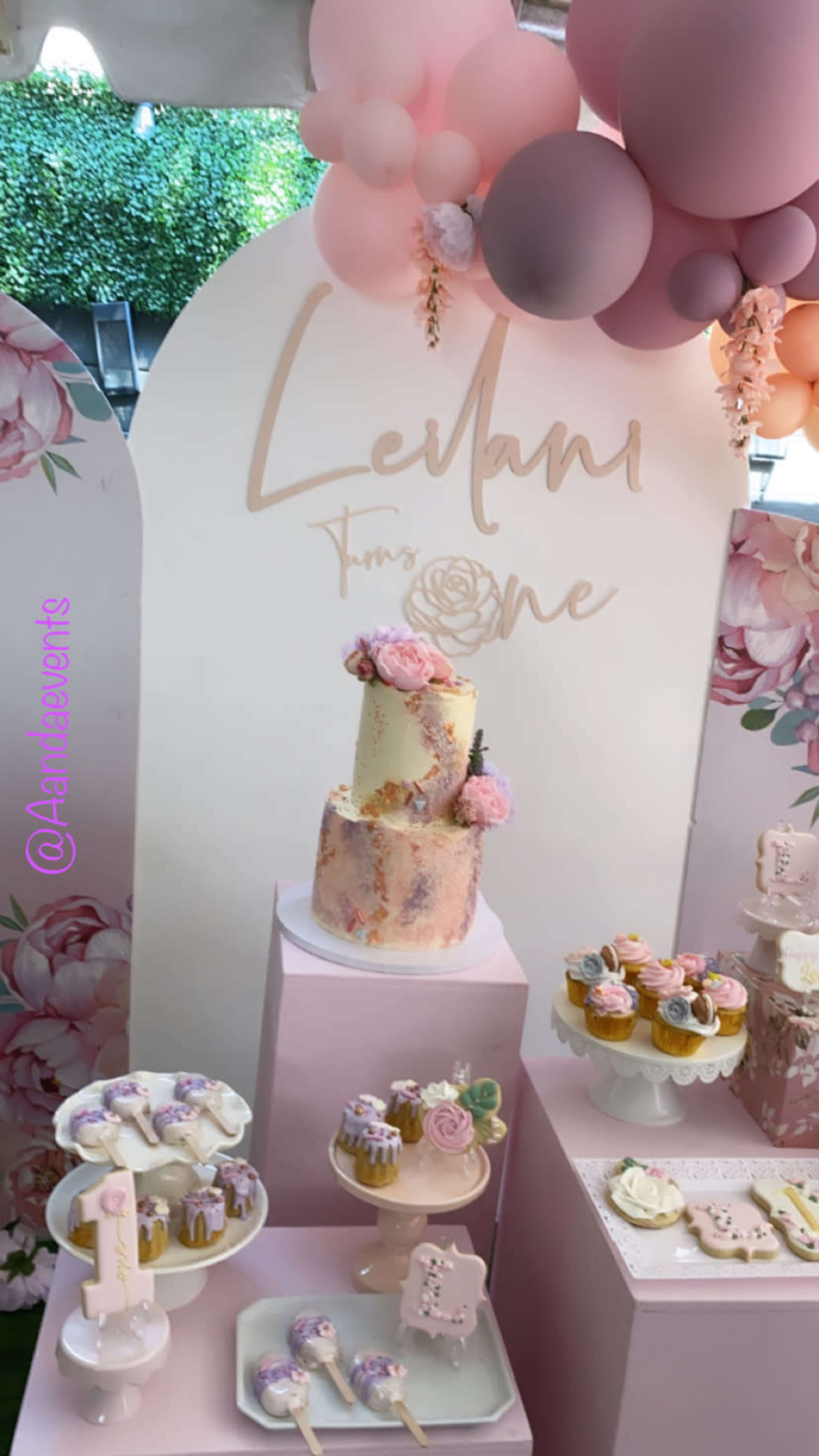 A decorative dessert display for a celebration, including a multi-tiered cake, various cupcakes, and cookies, set against a backdrop of pastel balloons and floral designs.