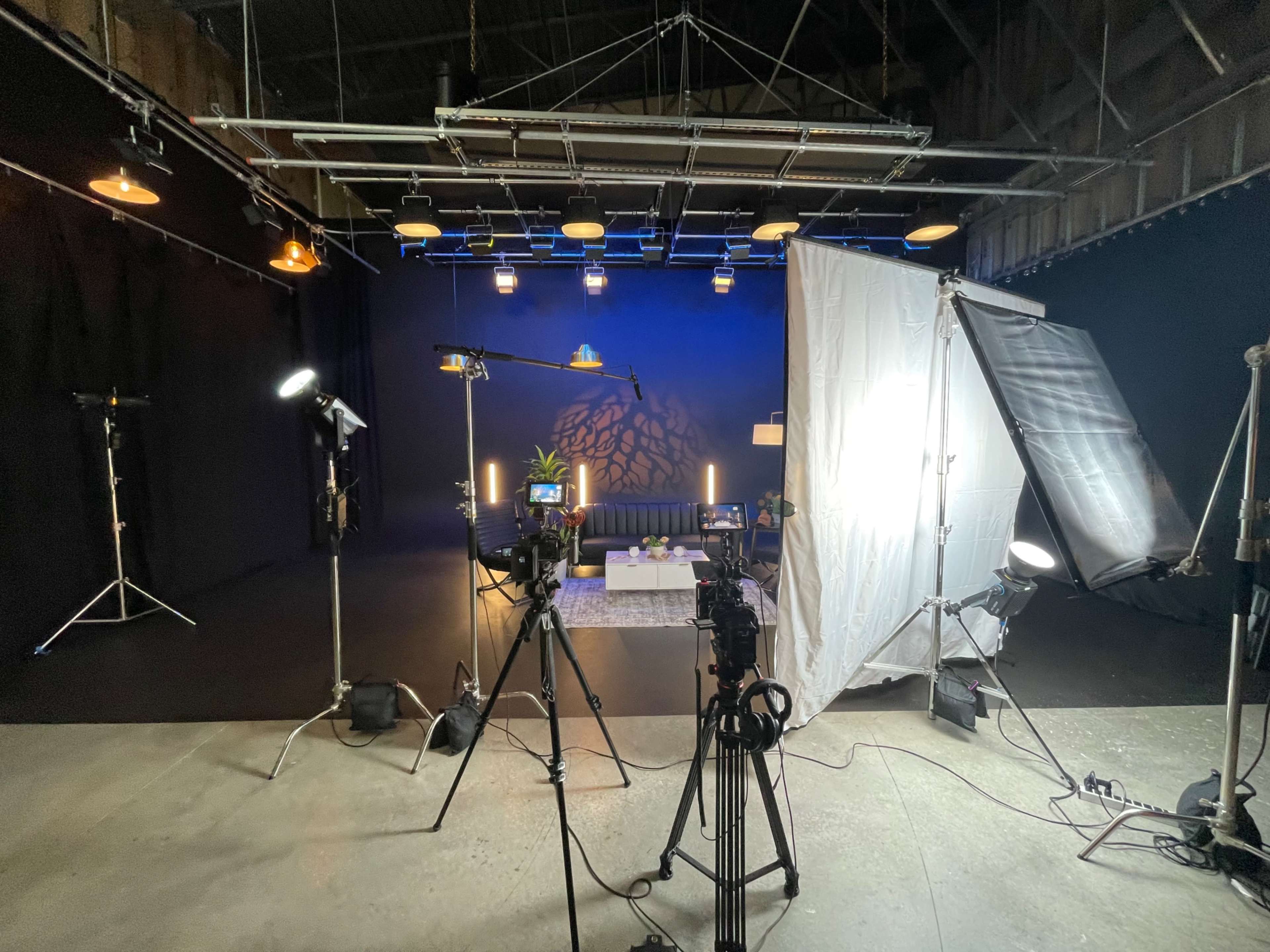 The image shows a dimly lit studio setup with multiple cameras, lighting equipment, and a small stage featuring a sofa and decorative elements.