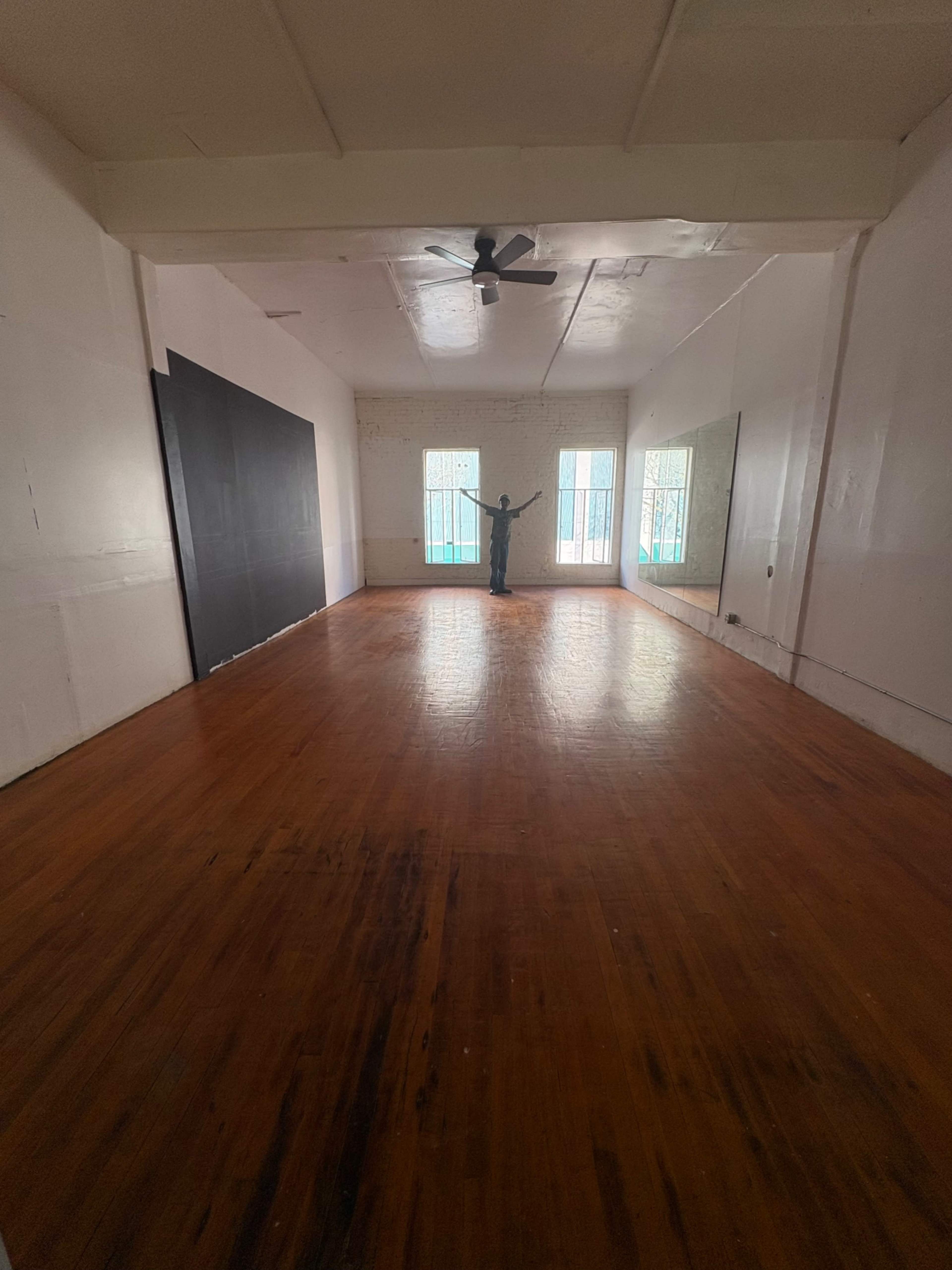 The image shows a spacious, empty room with hardwood floors, two windows letting in light, and a person standing in the center.