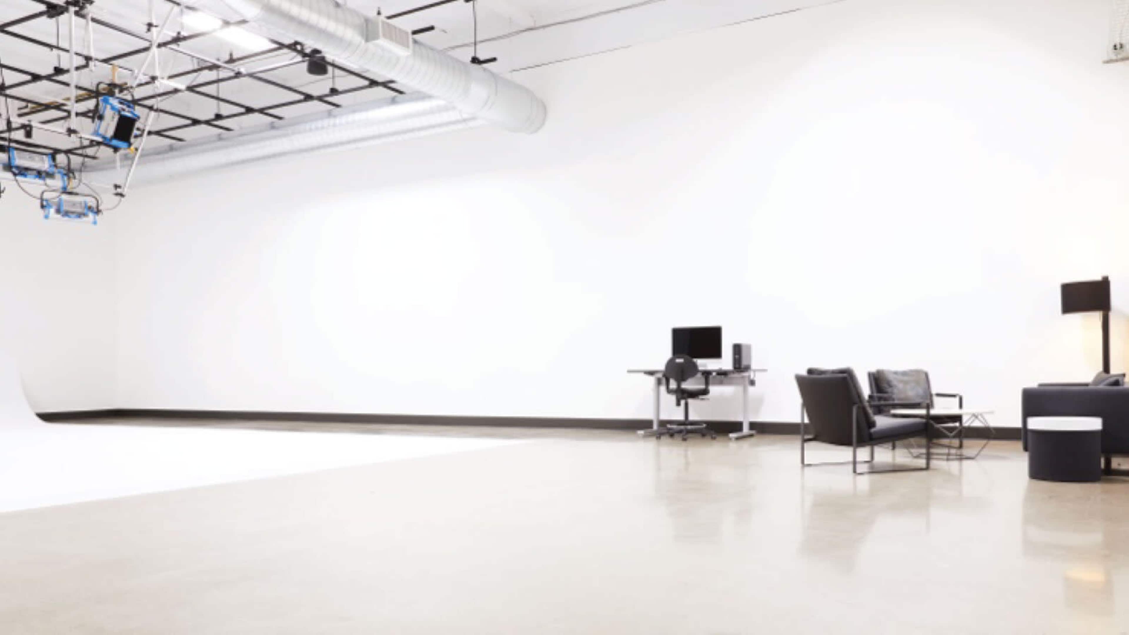 The image shows a spacious, empty studio with a white wall, a polished concrete floor, a desk with a computer, and a seating area featuring chairs and a table.