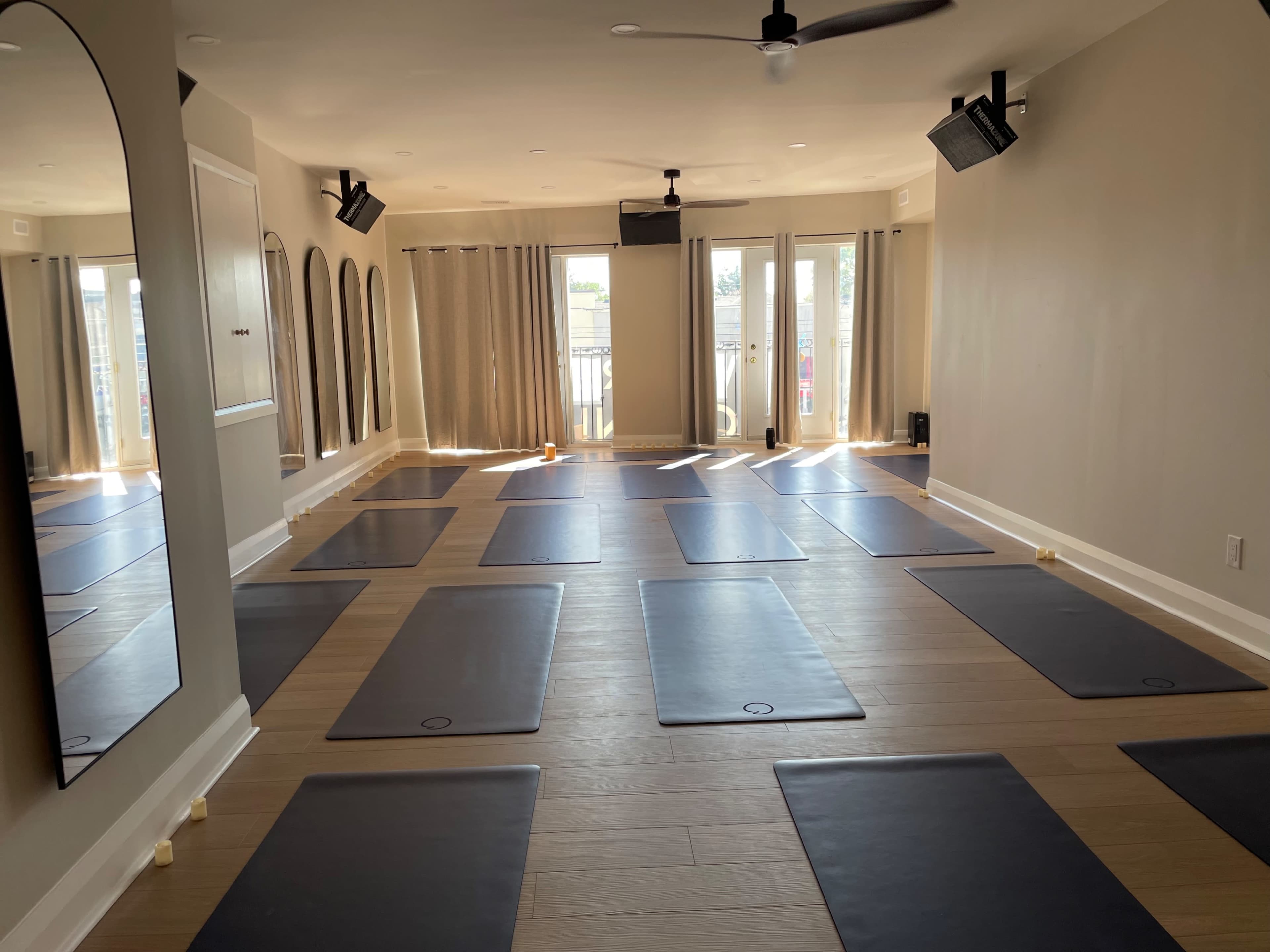 An empty yoga studio featuring several black mats arranged on a wooden floor, with large windows allowing natural light to enter.