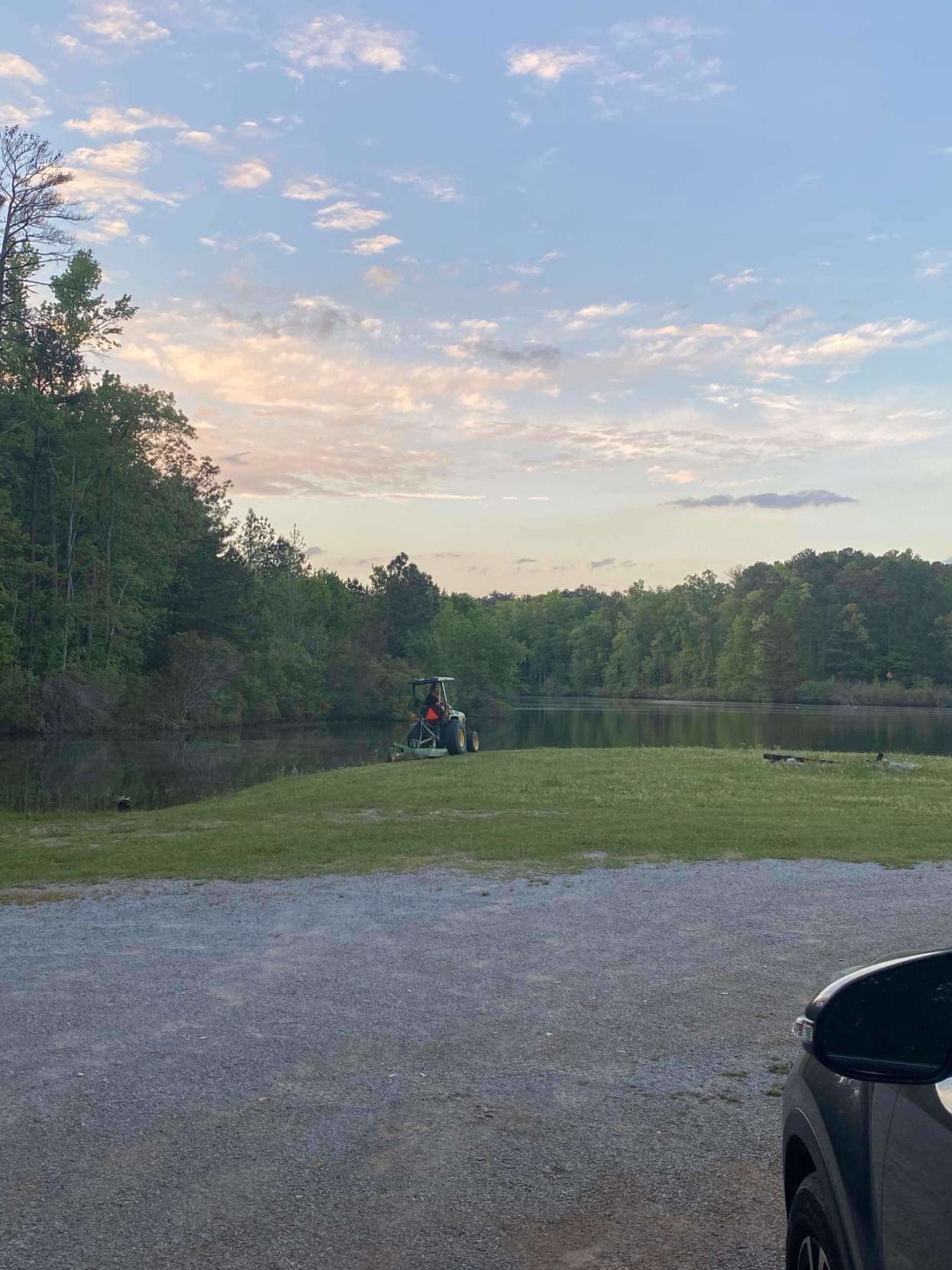 A tractor is parked at the edge of a grassy area beside a calm pond, with trees lining the background under a partly cloudy sky.