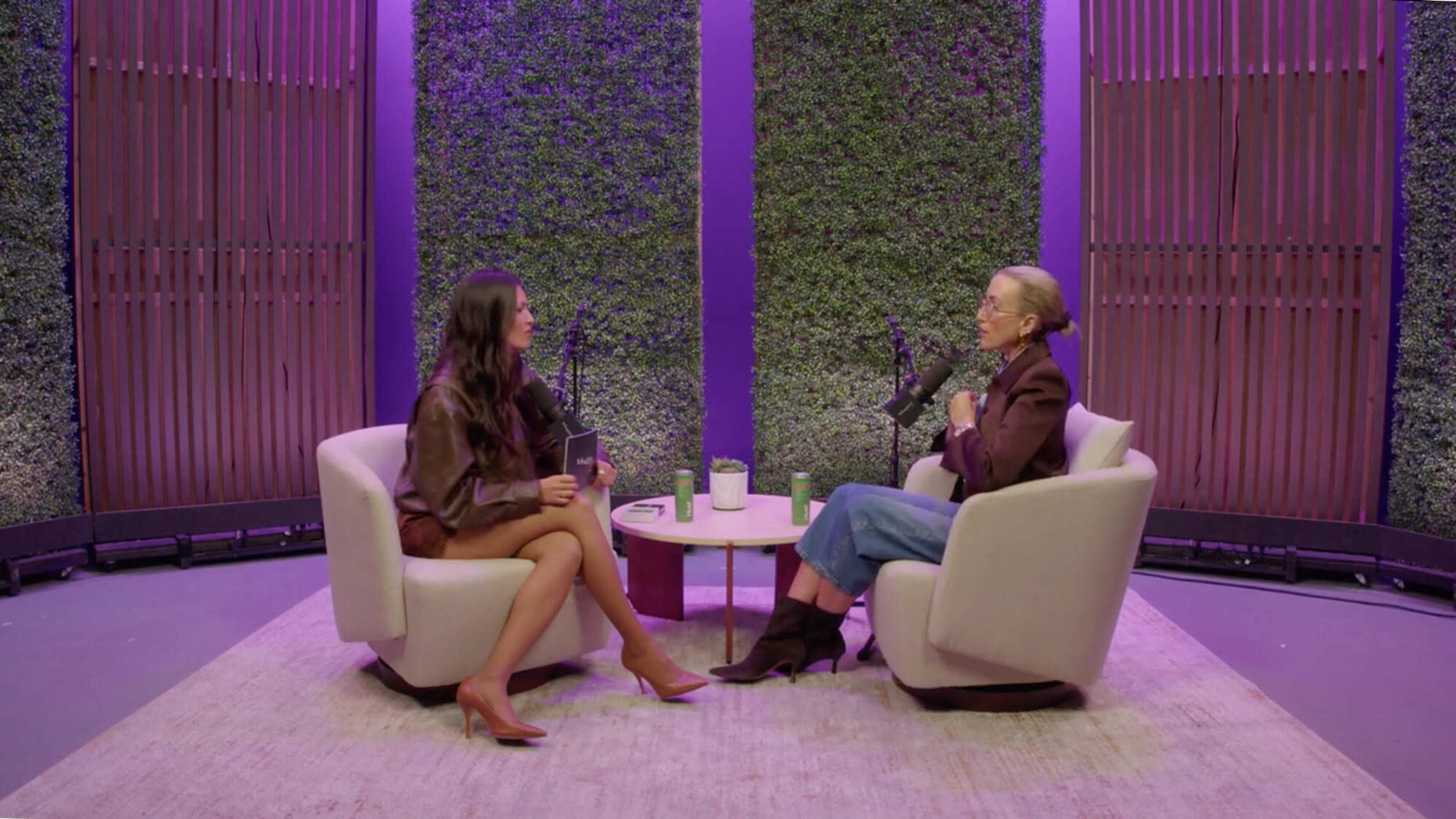 Two women sit in modern chairs during an interview, with a green wall covered in plants behind them.