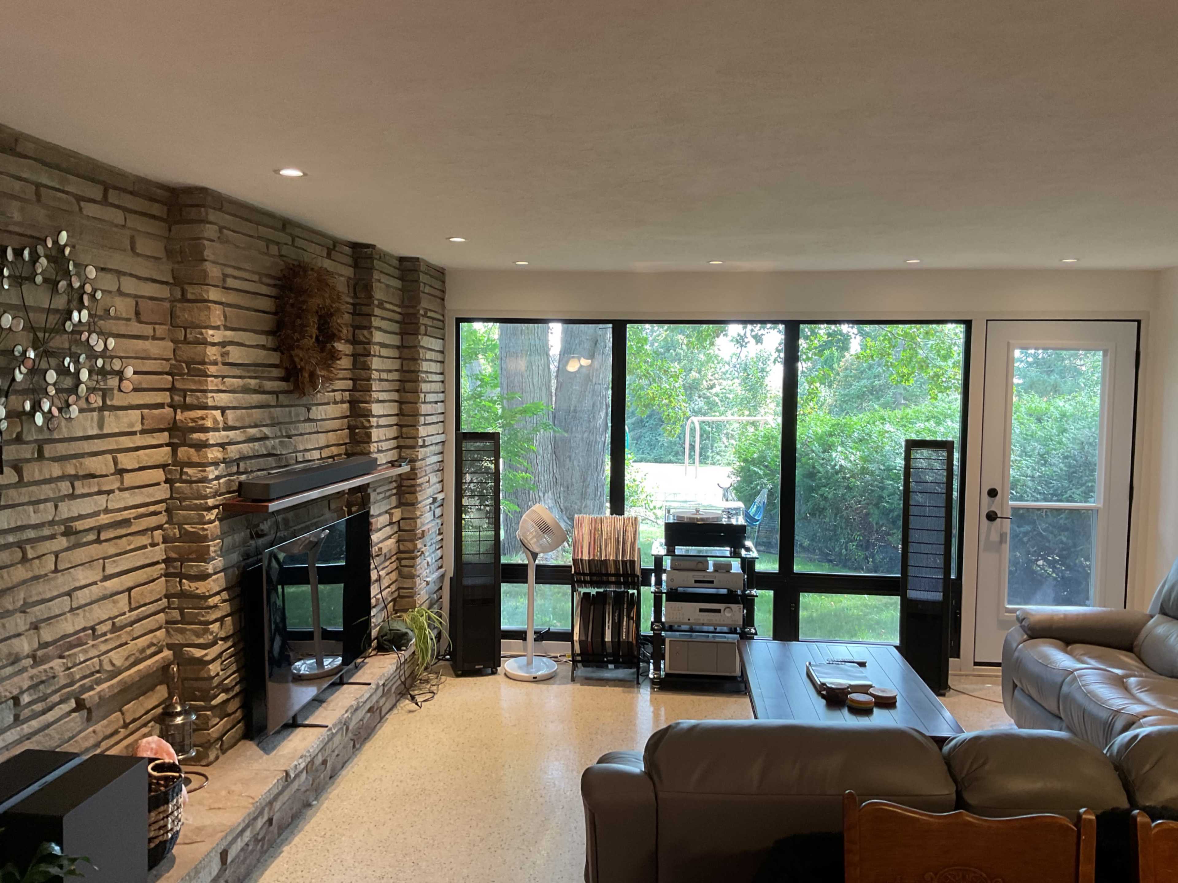 The image shows a cozy living room with a stone fireplace, a seating area, and large windows overlooking a green garden.