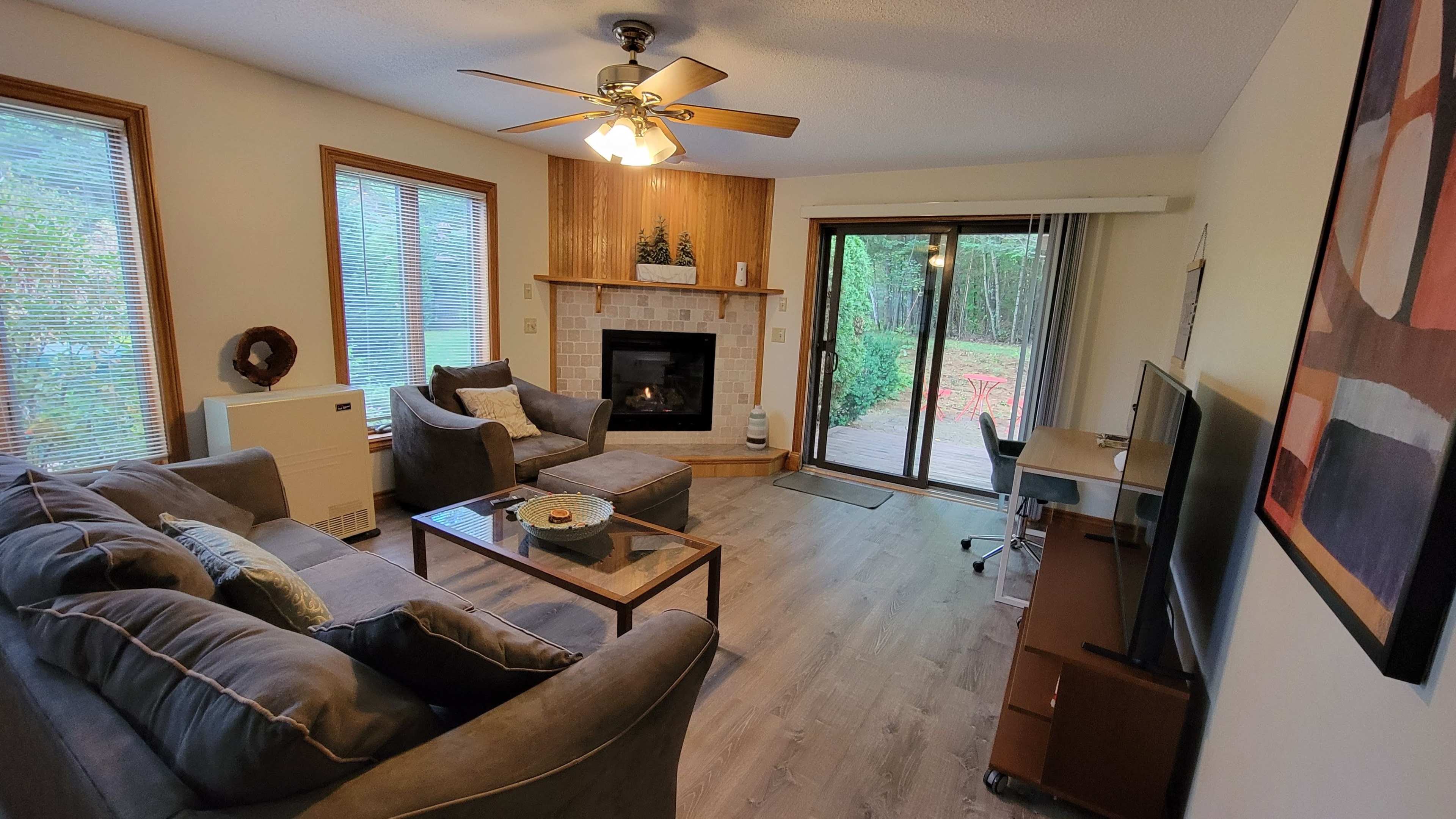 A cozy living room with a sectional sofa, a coffee table, a television, and a sliding glass door leading to a patio surrounded by trees.