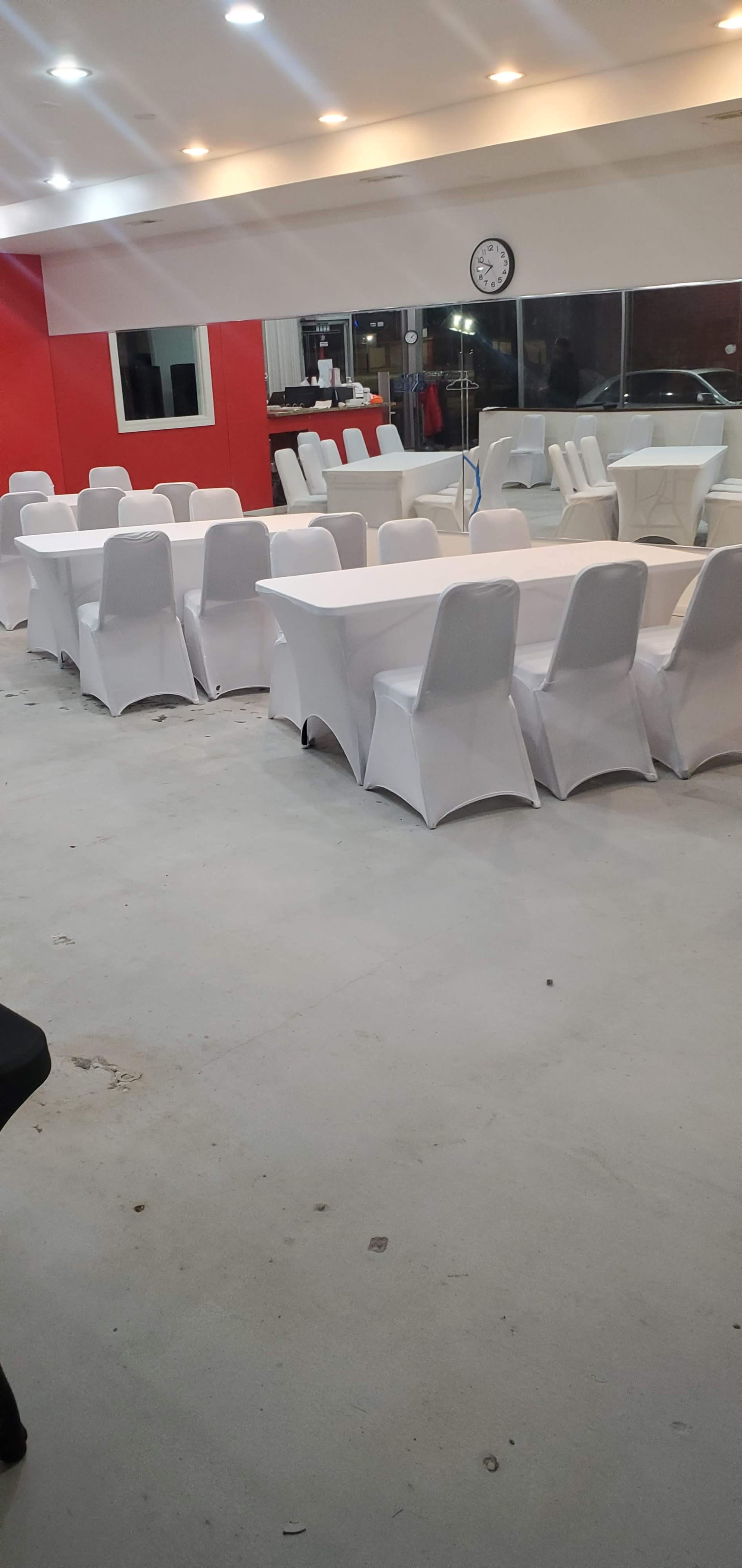 The image shows a spacious room with several tables covered in white tablecloths and surrounded by white chairs, against a red wall.