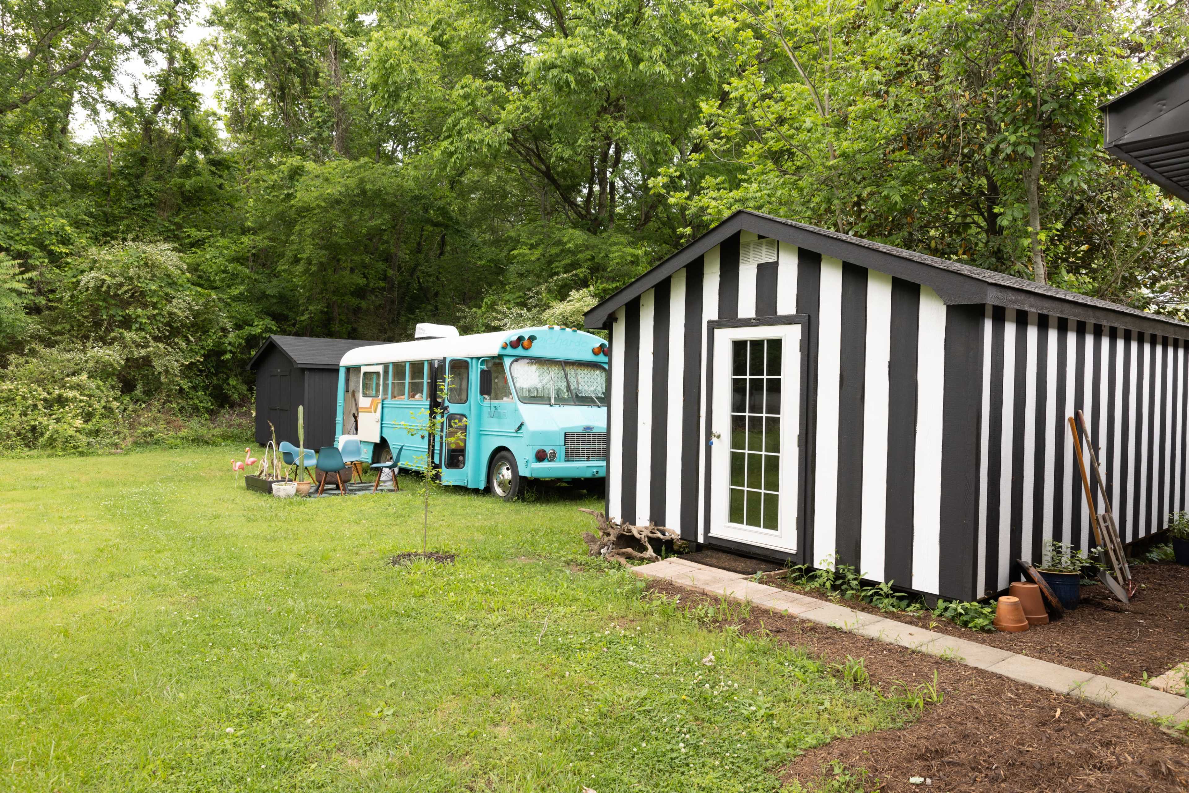 A turquoise bus is parked beside a striped black-and-white shed in a green yard surrounded by trees.
