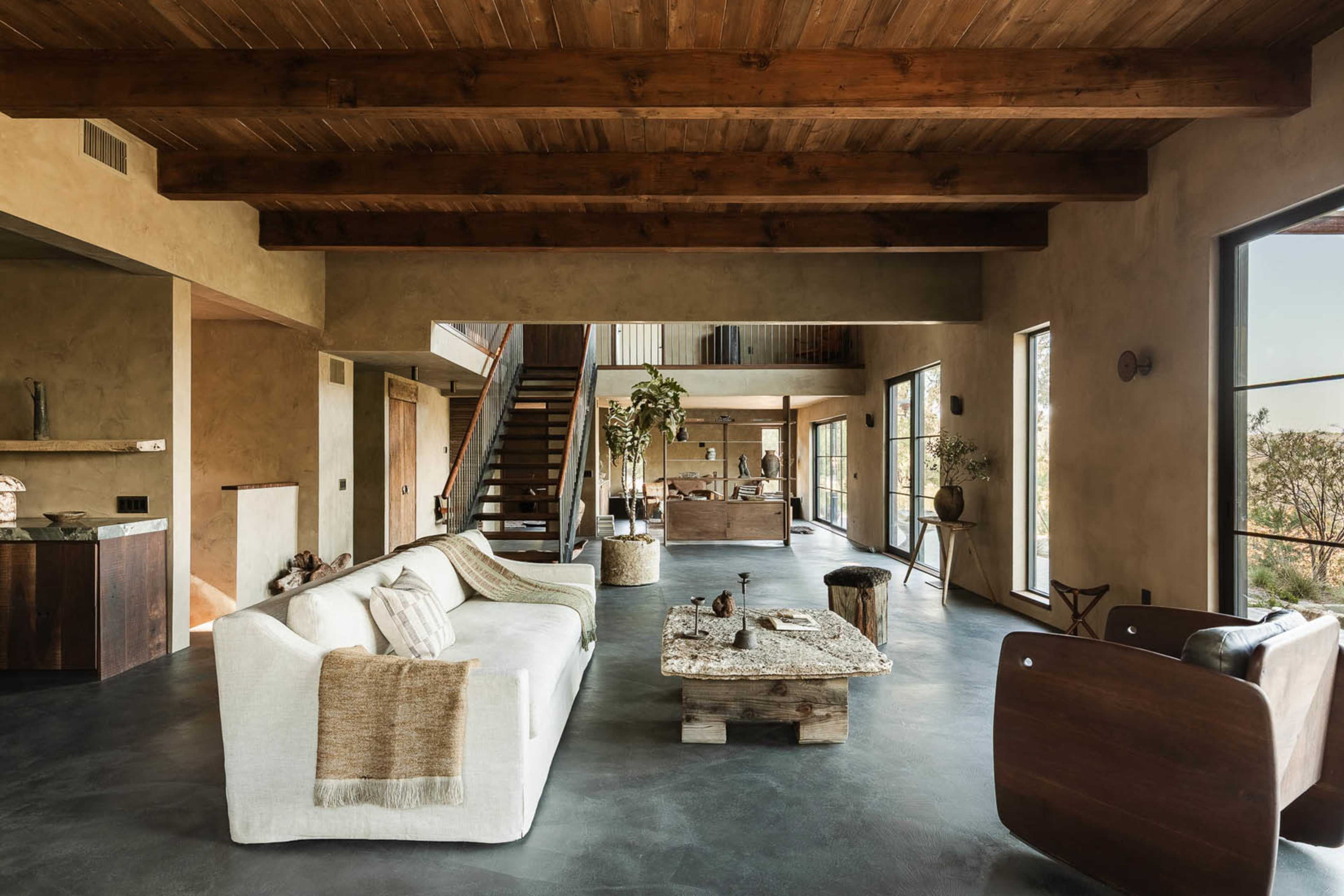 The image shows a spacious, modern living area with a white sofa, a wooden coffee table, and large windows that allow natural light to enter.