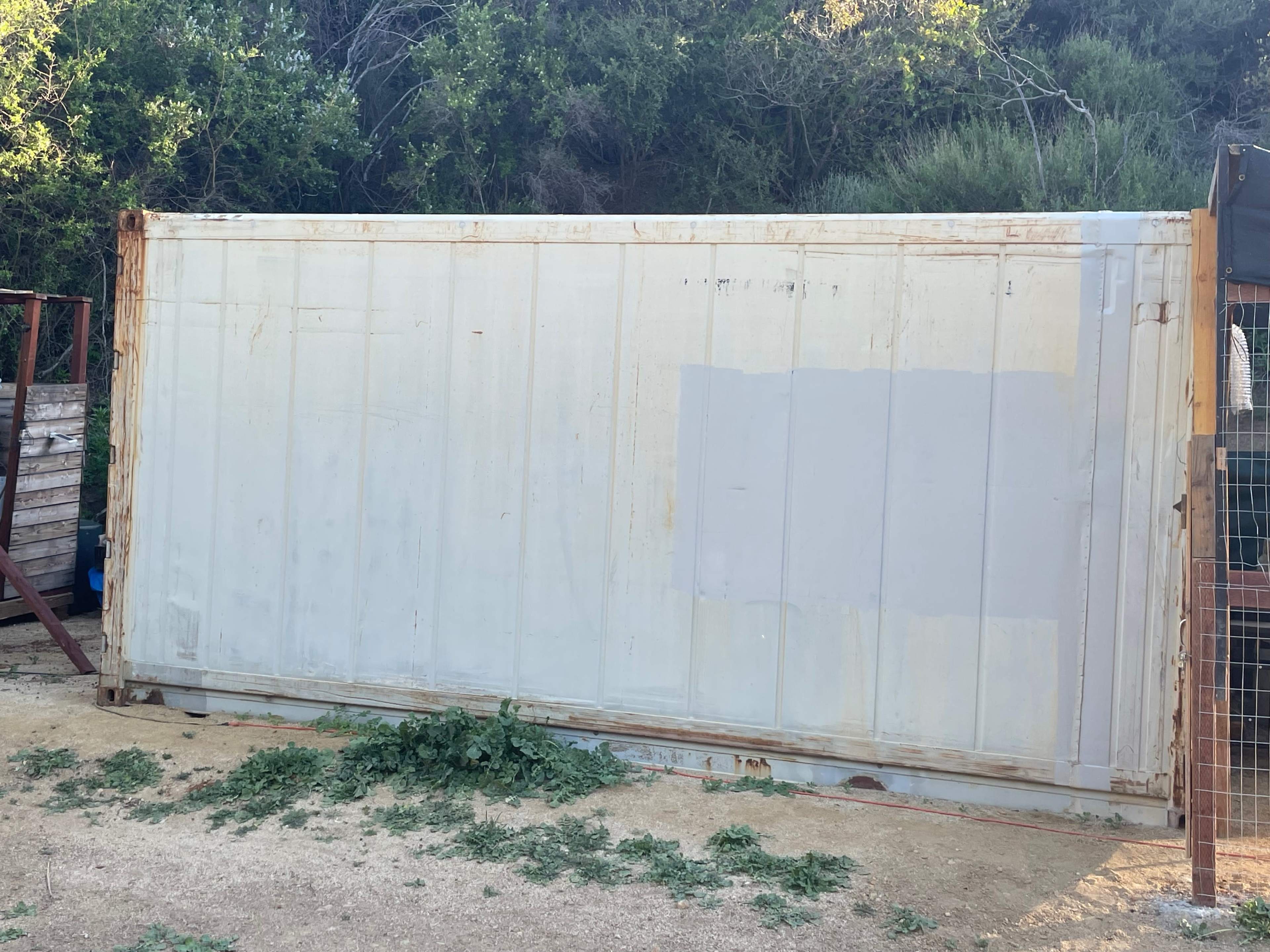 A weathered shipping container stands on a dirt surface, partially covered with greenery and showing signs of age.