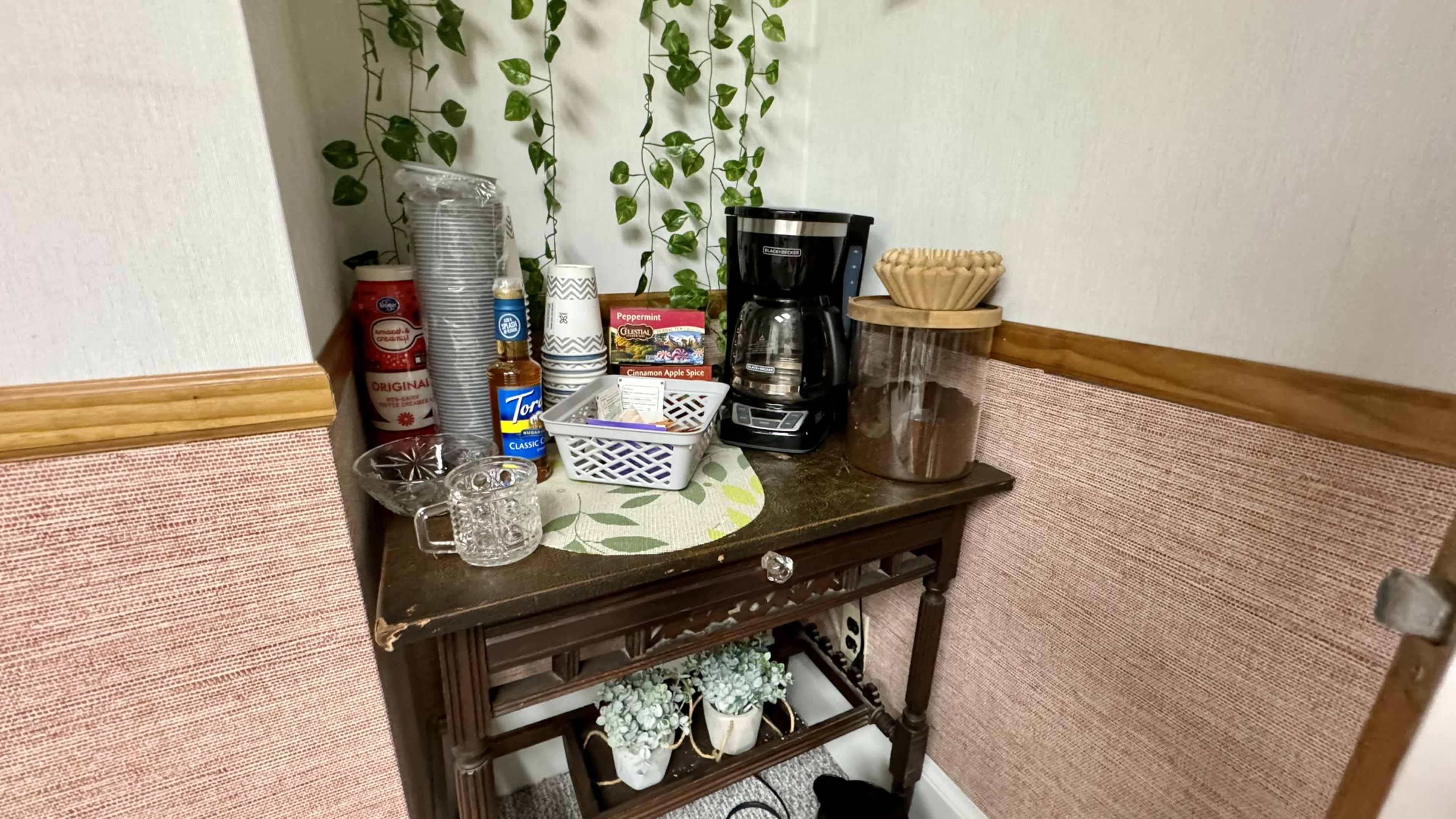 The image shows a corner table with a coffee maker, several containers, and glasses, surrounded by green plants on the wall.