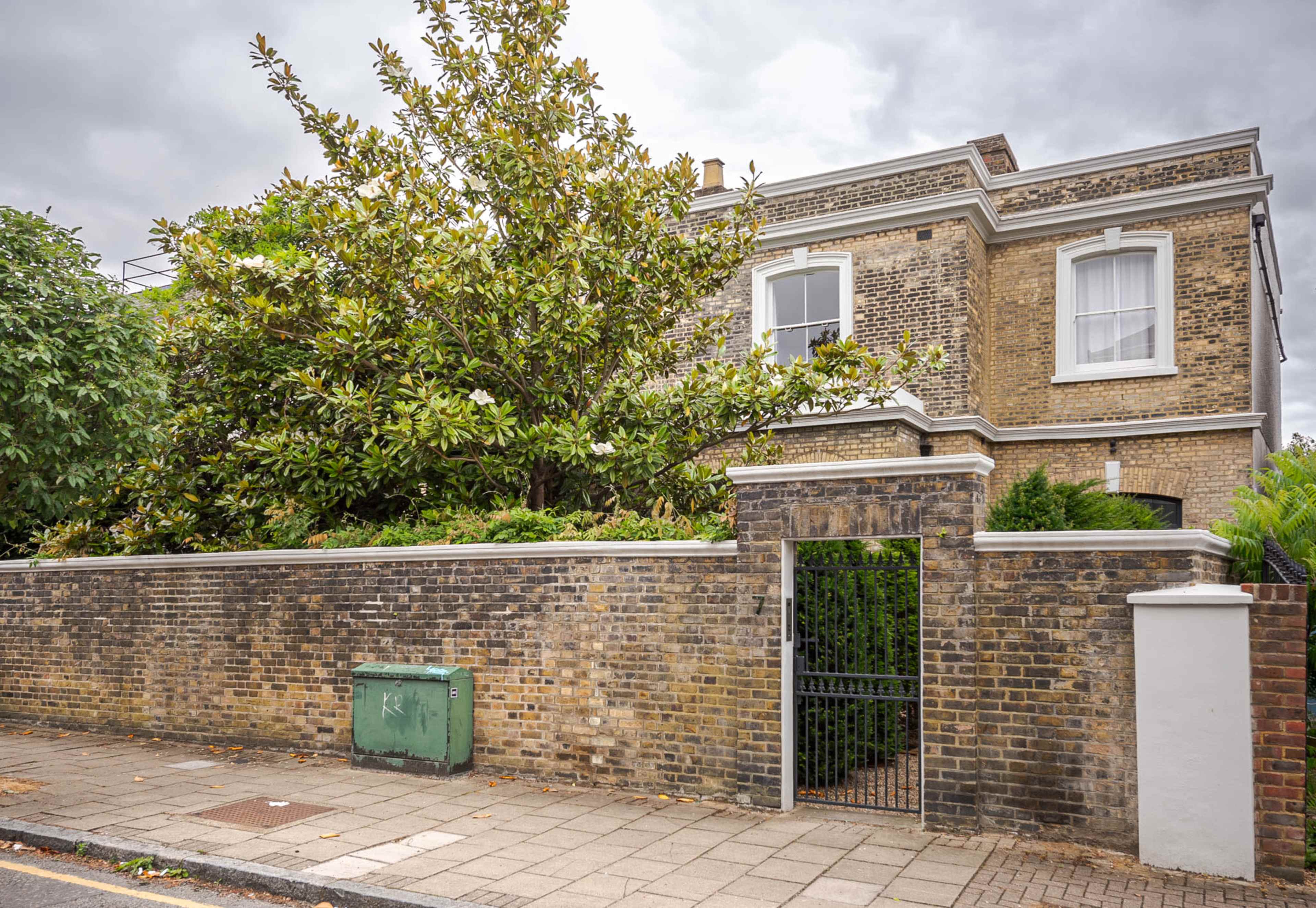A two-story brick house with a large tree in the yard is situated behind a low brick wall and gate.