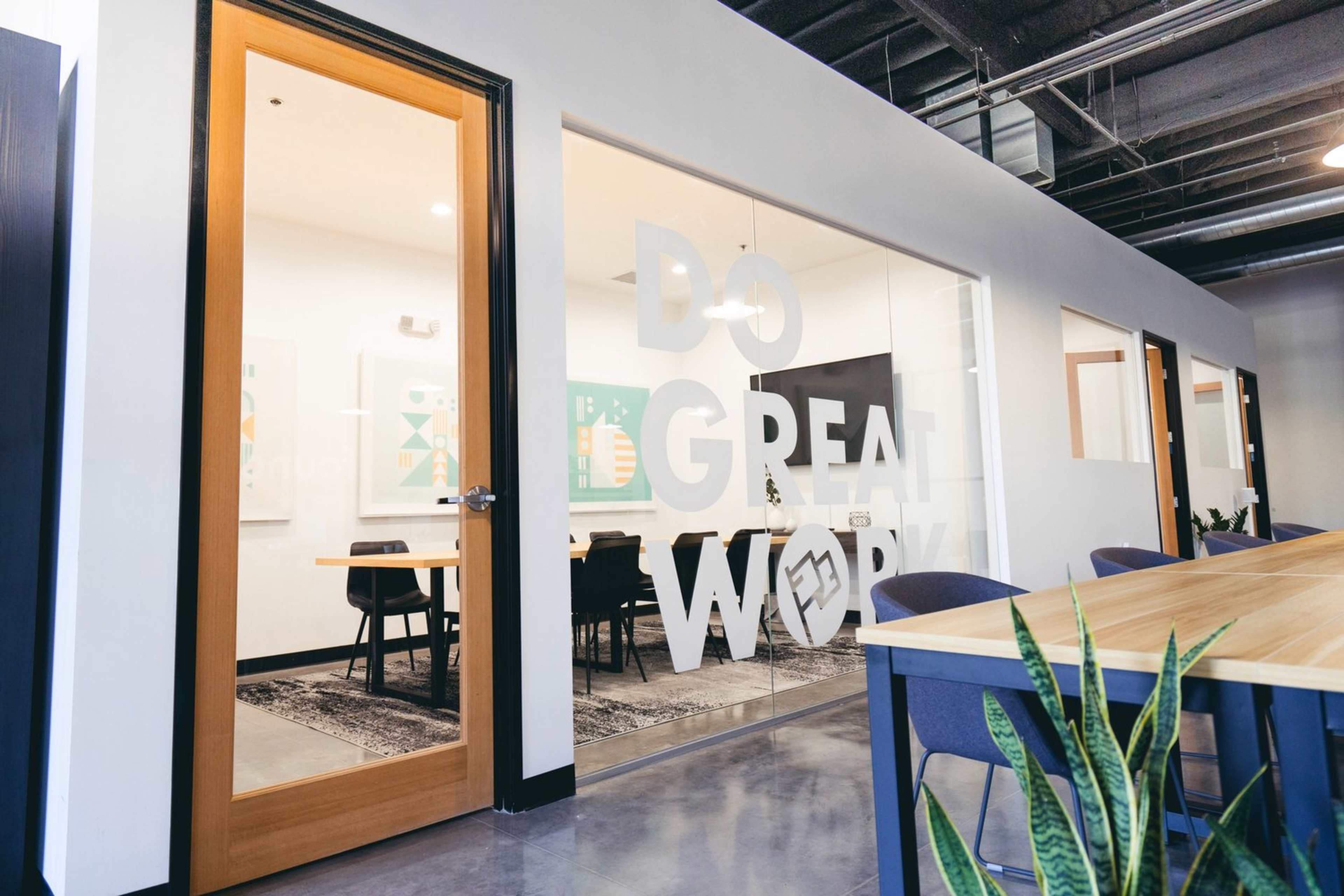 A modern office space with a glass-walled meeting room featuring the phrase "DO GREAT WORK" and a large wooden table surrounded by chairs.