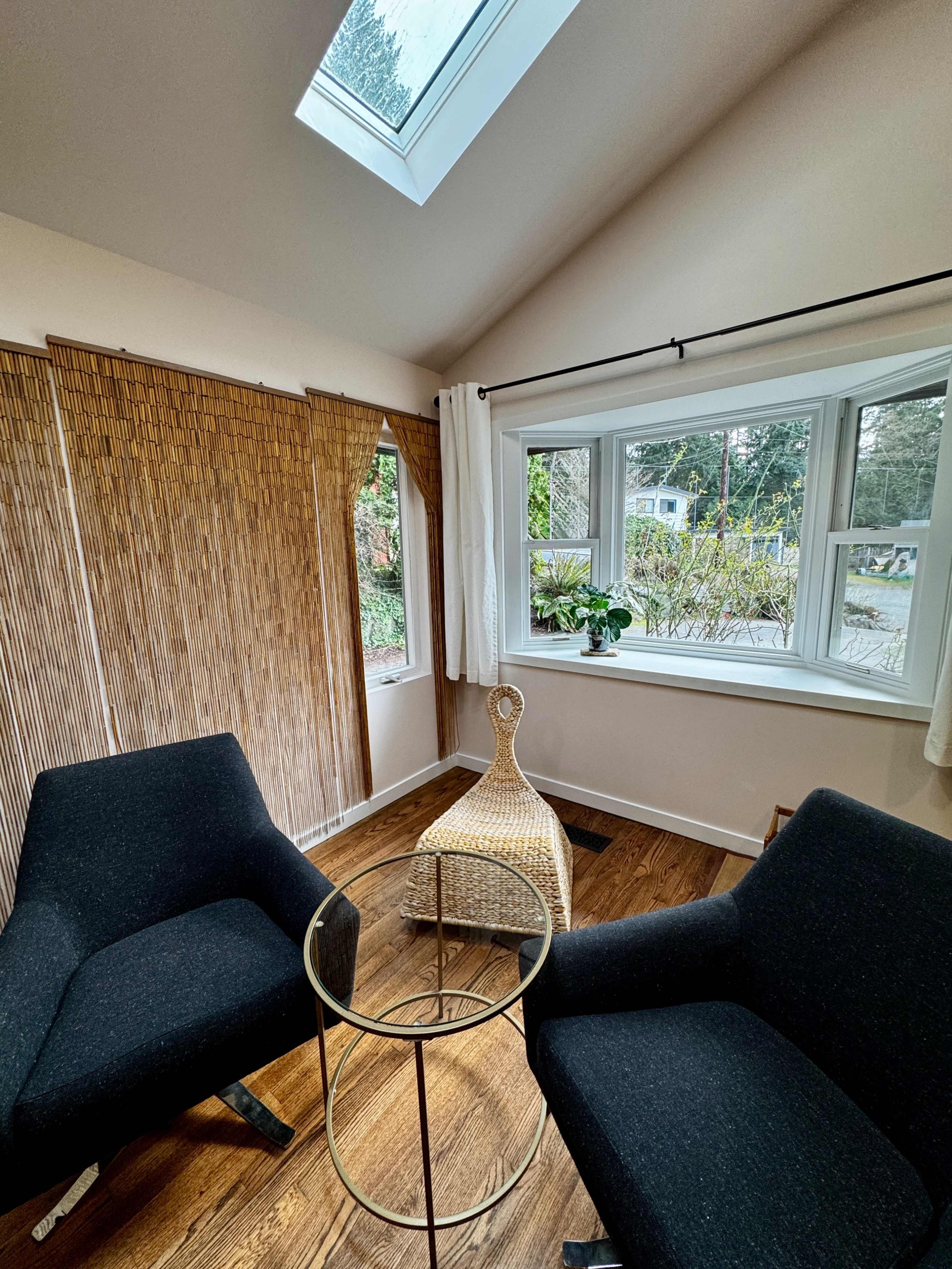 The image shows a cozy corner of a room featuring two black armchairs, a round glass side table, and natural light streaming in through a window with a view of greenery outside.