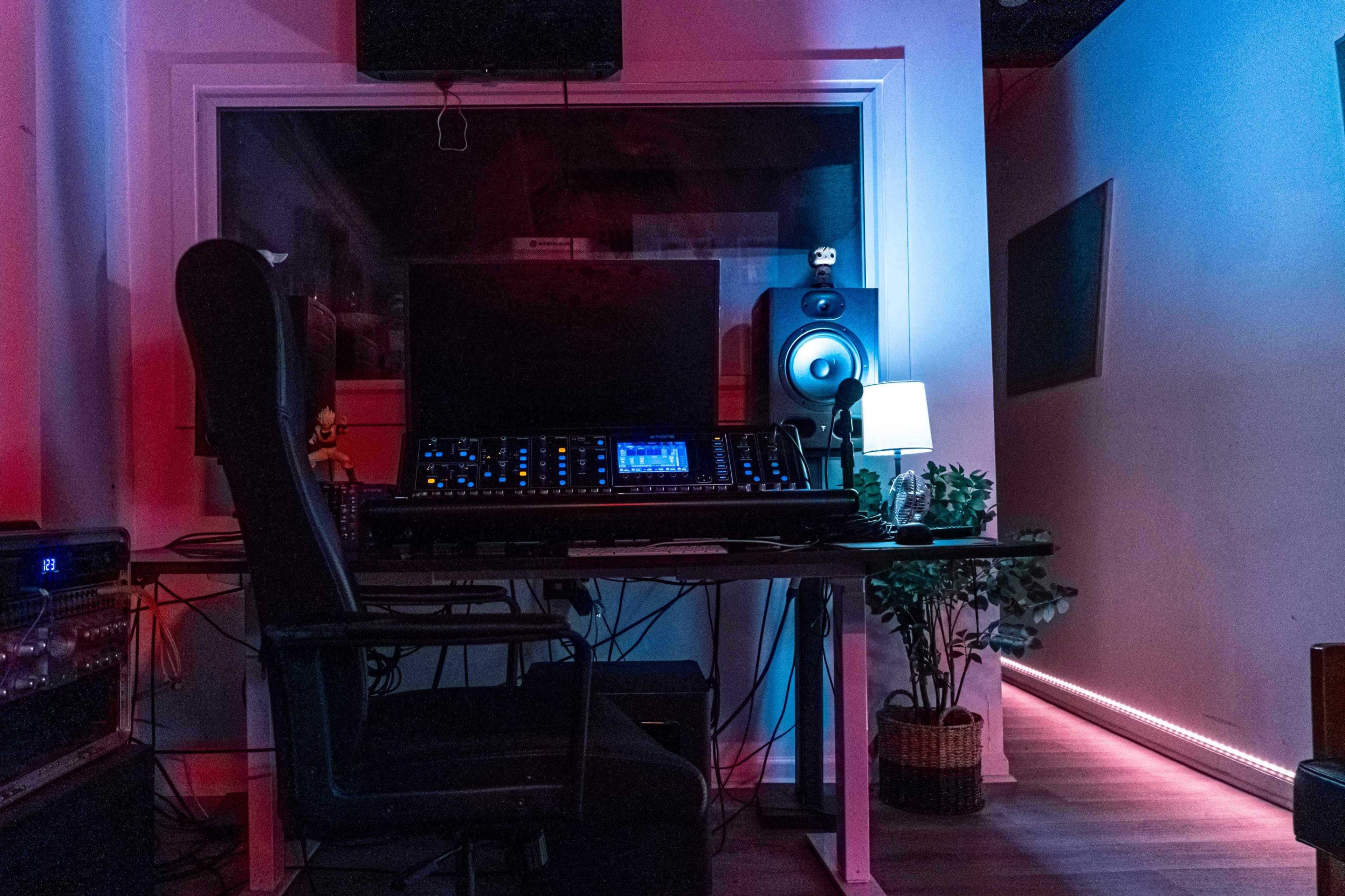 A music studio with a desk featuring audio equipment, a chair, and a small lamp, illuminated by colored lights.