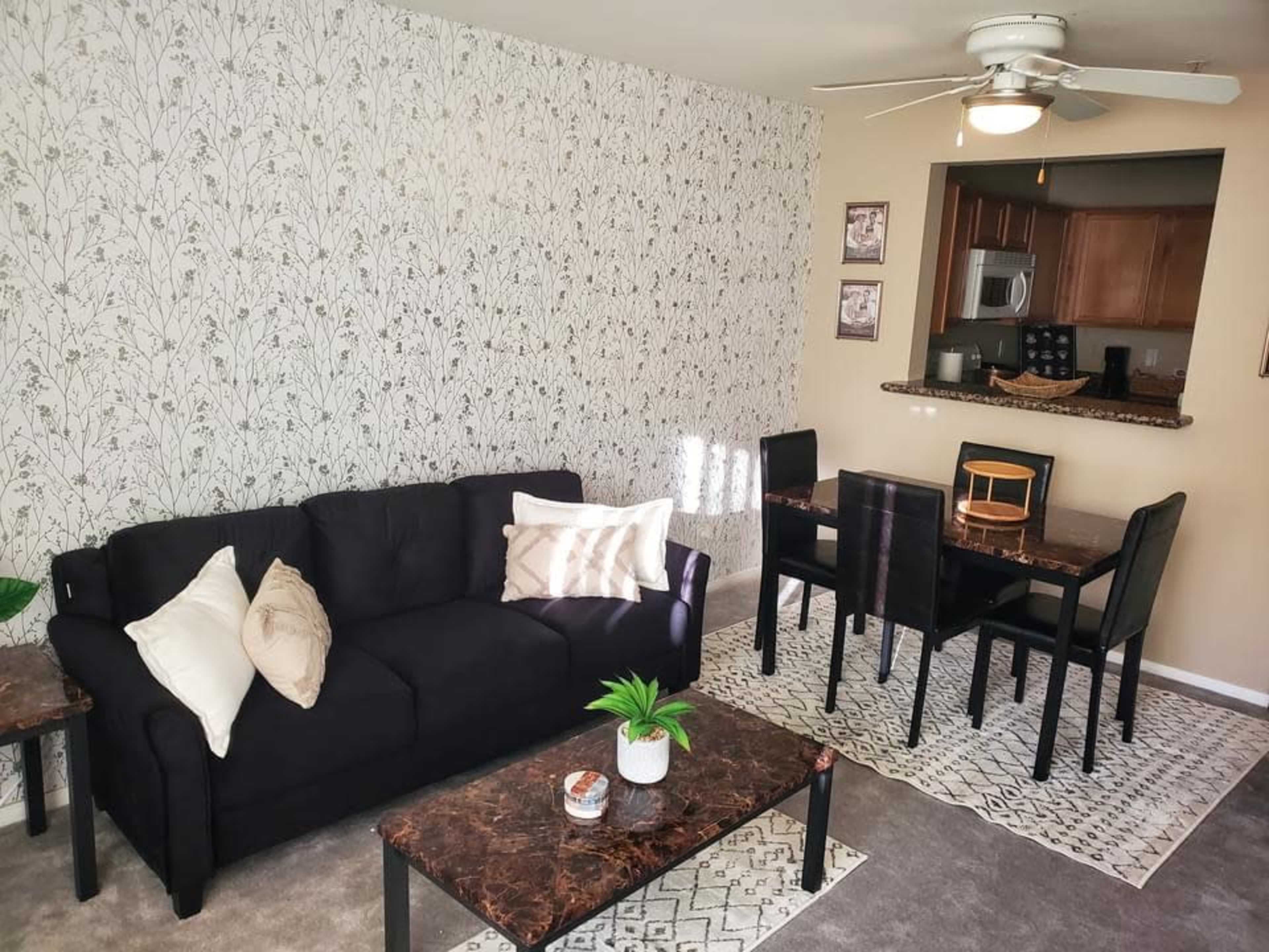 A living room with a black sofa, patterned coffee table, and a dining area in the background featuring a round wooden table and black chairs.