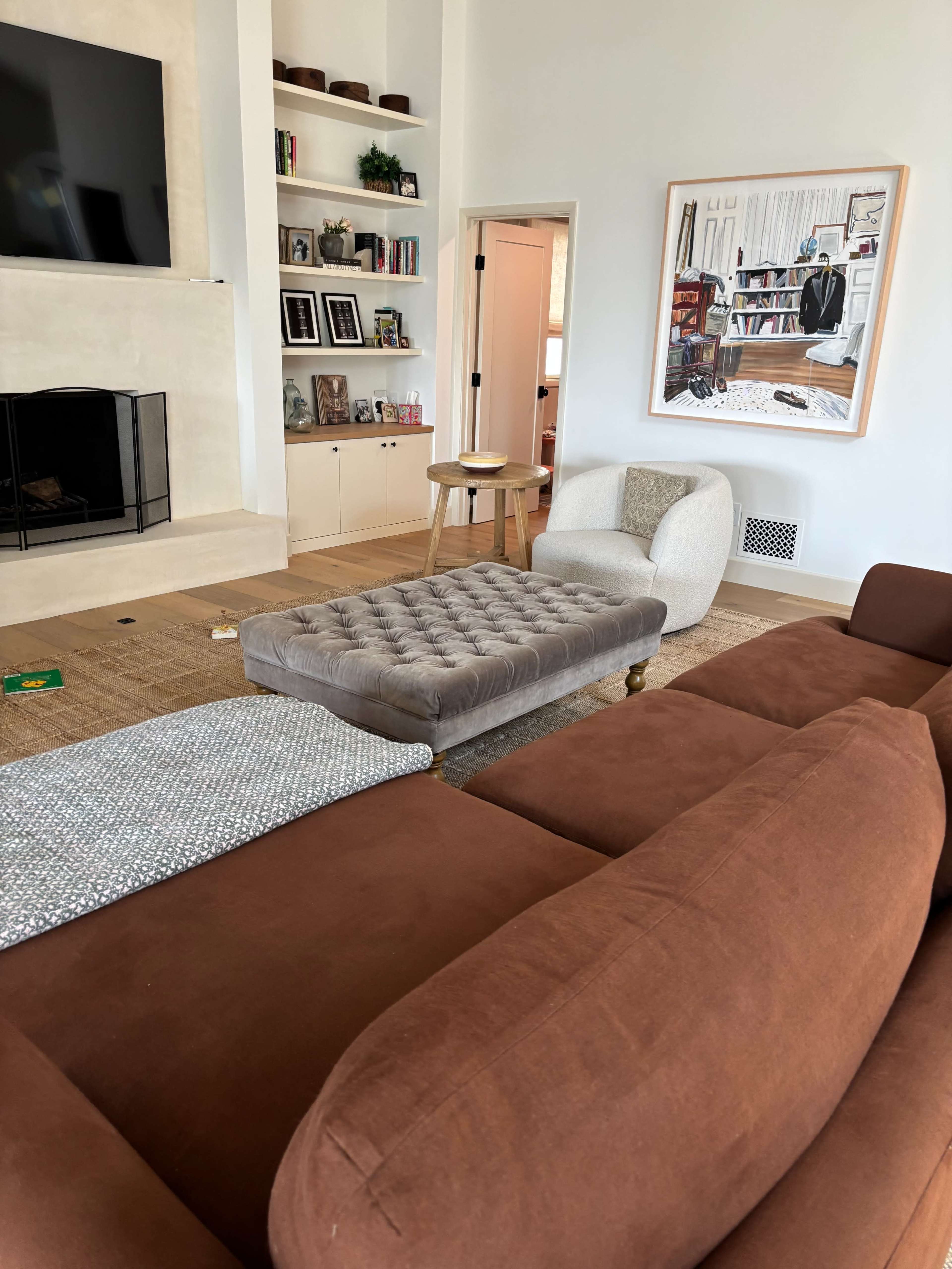 The image shows a living room with a brown sofa, a tufted ottoman, and a small wooden side table, along with shelves displaying books and decor.