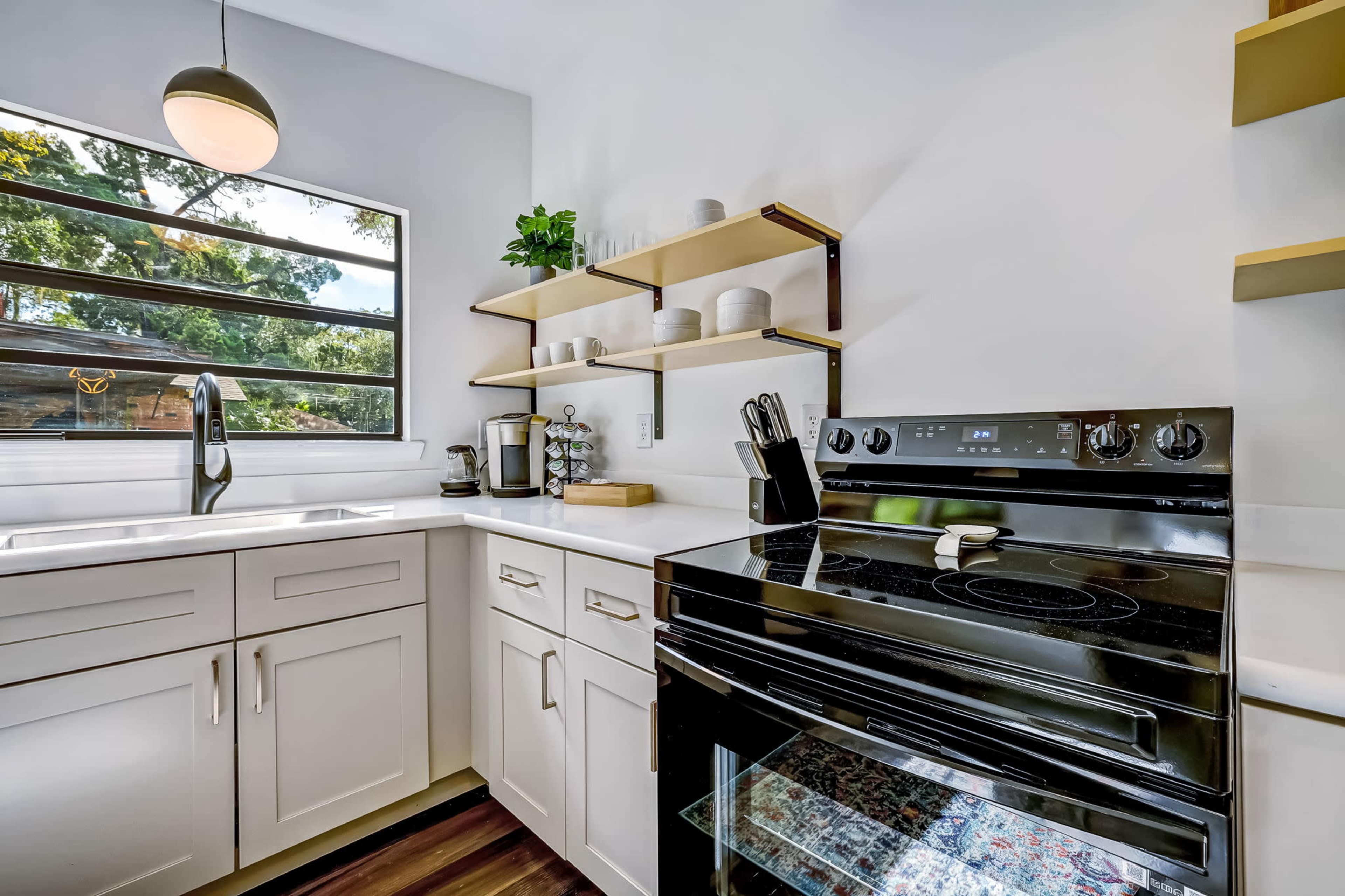 A modern kitchen with a black stove, white cabinets, open shelving, and a large window letting in natural light.