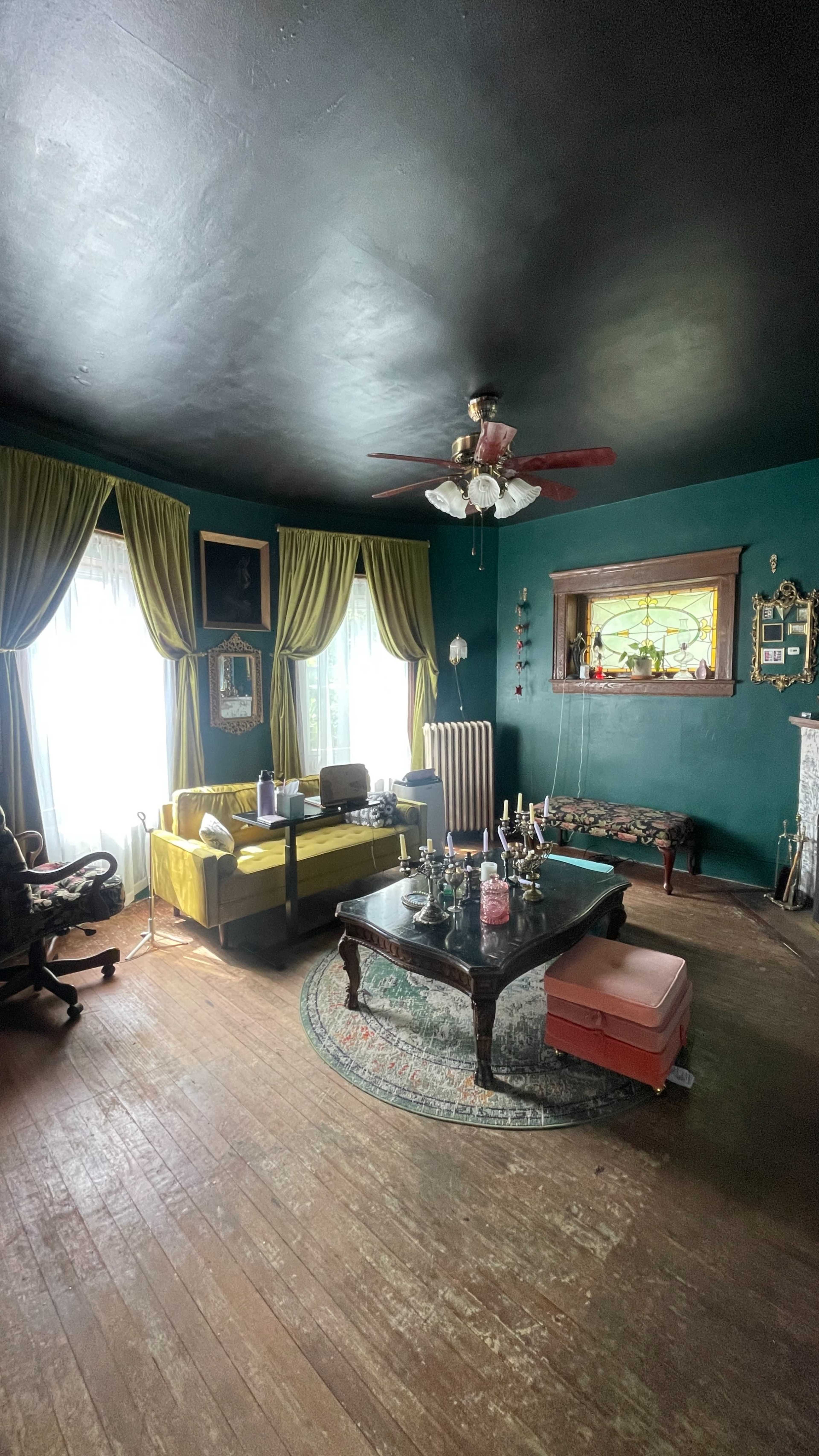 The room features a green wall, large windows with sheer white curtains, a yellow sofa, a wooden table, and various decorative items displayed on the walls and furniture.