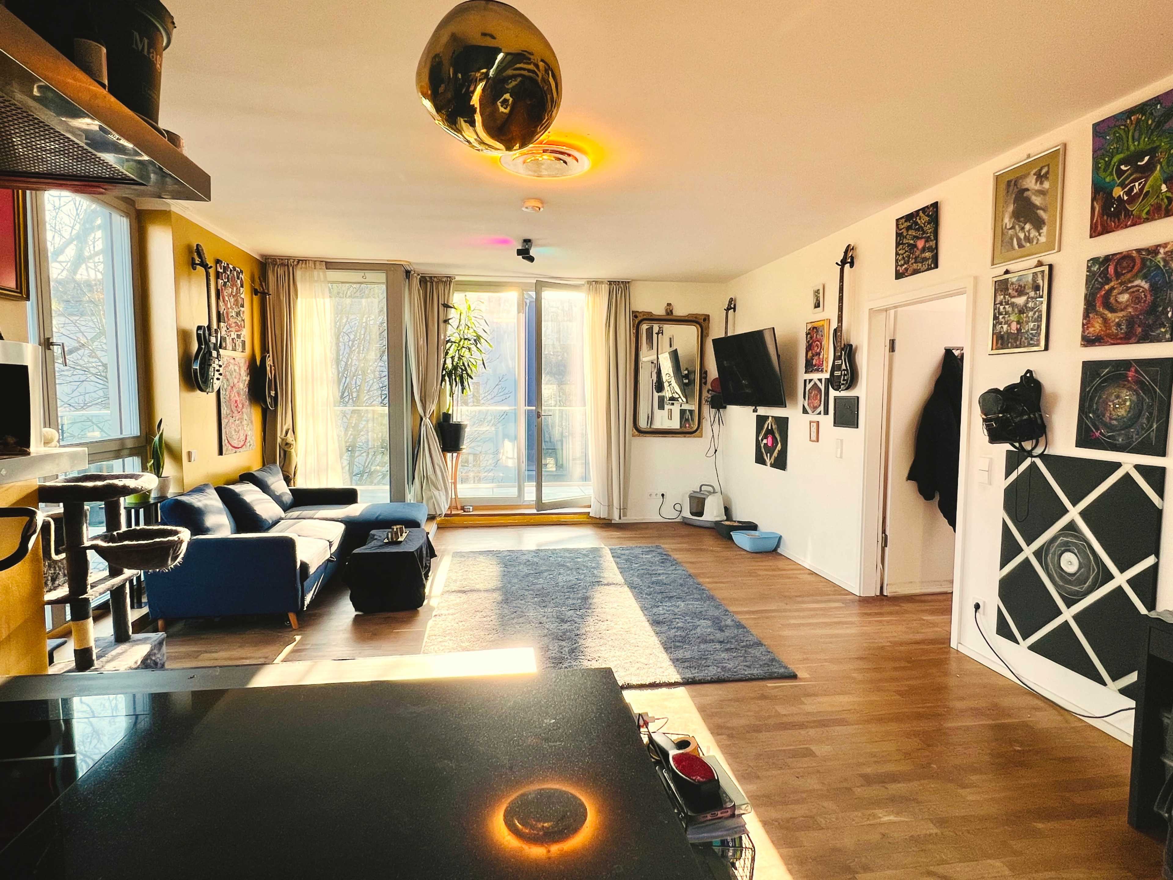 The image shows a spacious living area with a blue couch, large windows letting in natural light, and various artworks displayed on the walls.