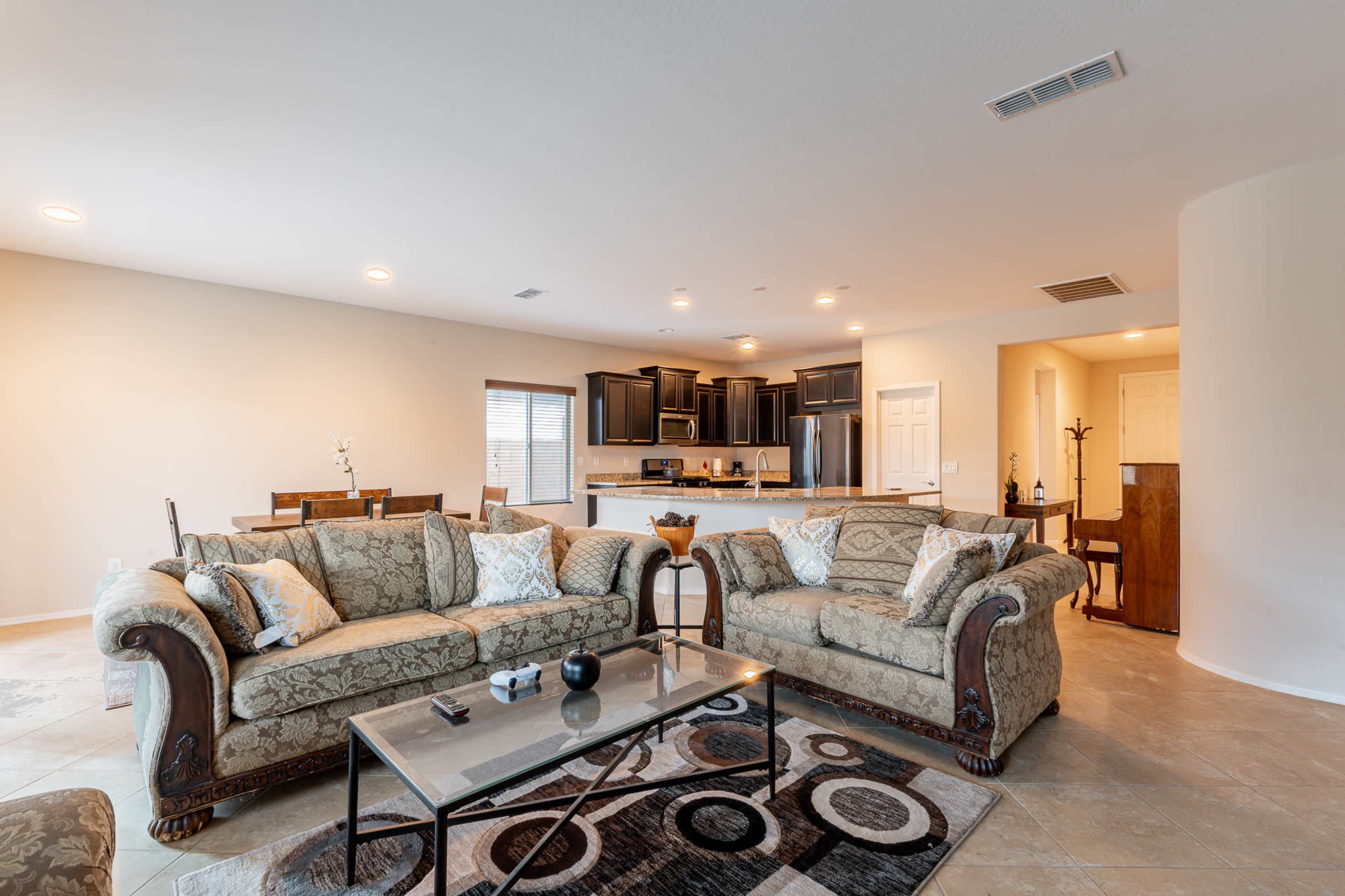 The image shows a spacious living room with two patterned couches facing a glass coffee table, leading into an open kitchen area with dark cabinetry.